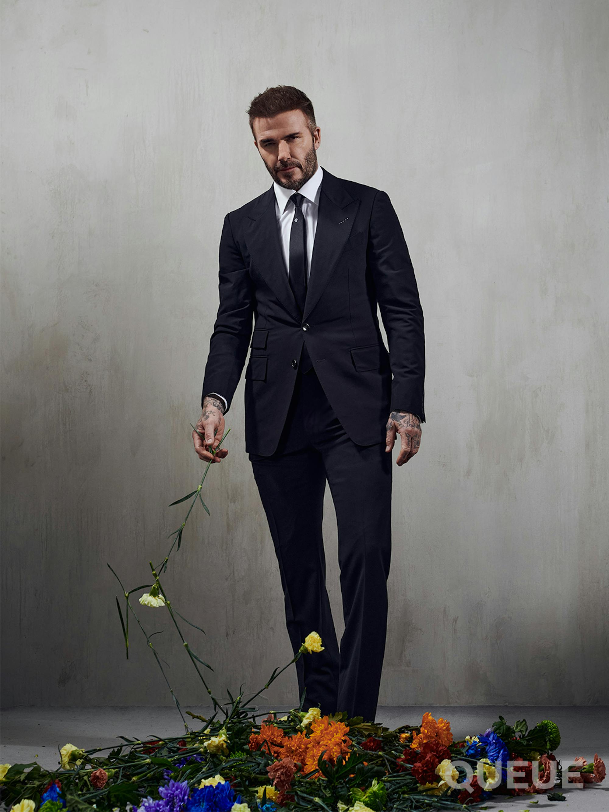 David Beckham wears a suit and walks through a pile of flowers in a gray-walled room.