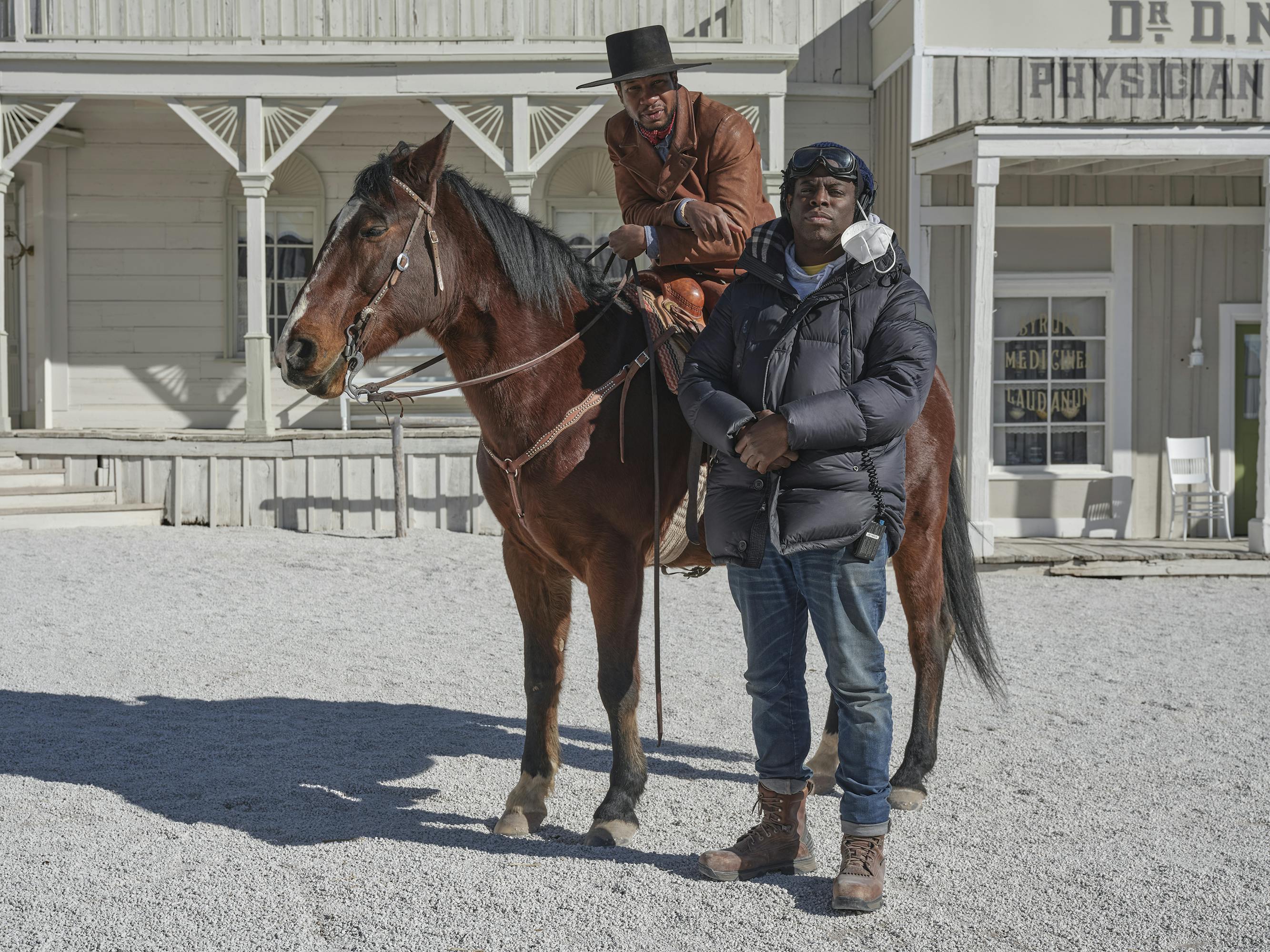 Jonathan Majors rides a brown horse and Jeymes Samuel stands next to him in a puffer jacket, jeans, and a black helmet. Behind them is the white town, complete with white sand and a white wraparound porch.