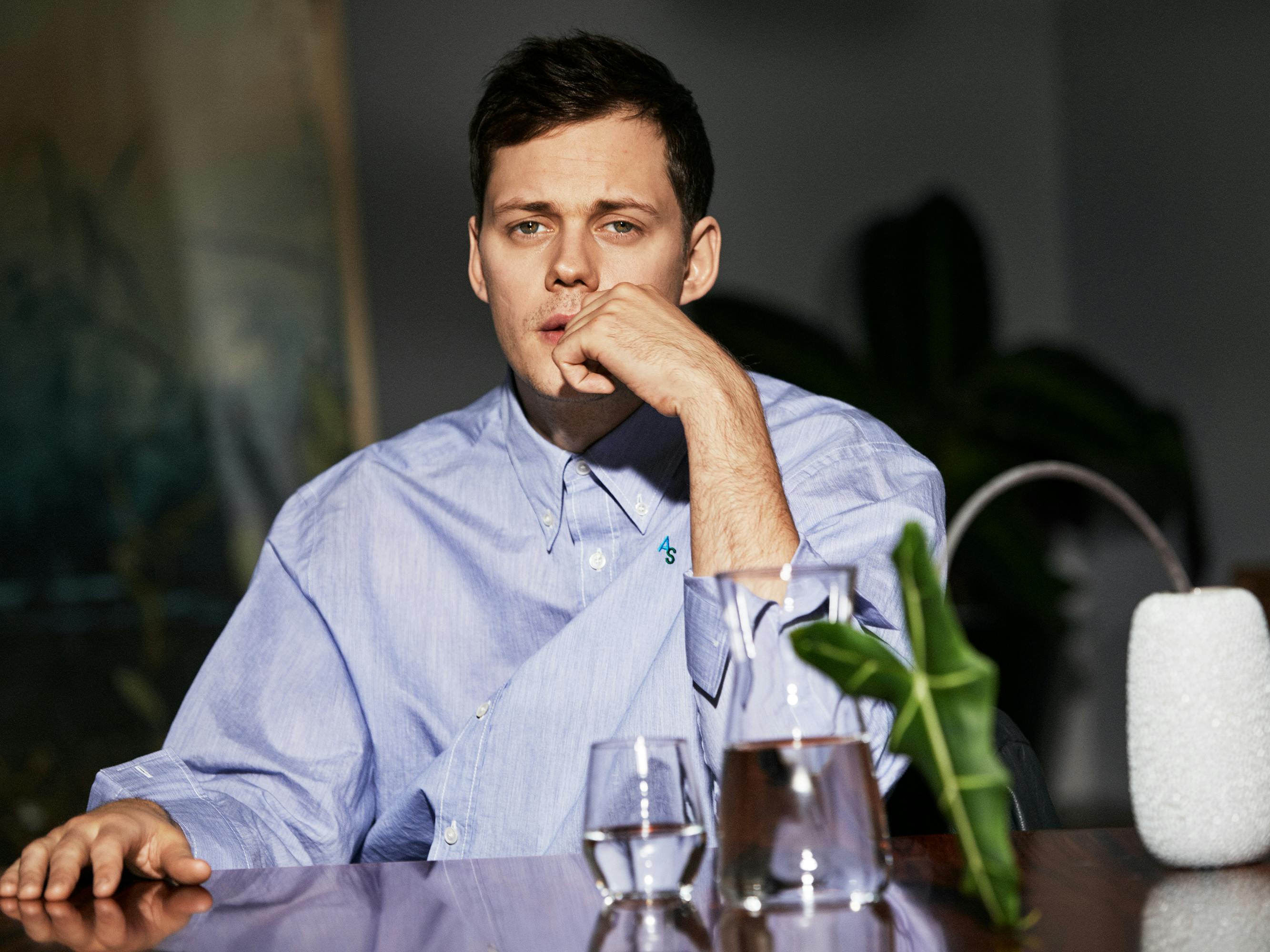 Bill Skarsgård wears a blue buttoned-down shirt and sits at a table strewn with glasses and a plant.