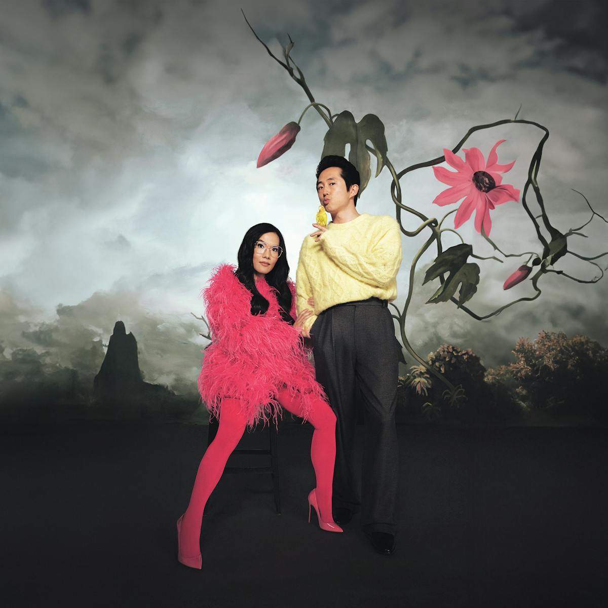 Ali Wong and Steven Yeun pose together against a dramatic cloudy background. Wong wears a pink ensemble; Yeun wears charcoal pants and a yellow sweater.