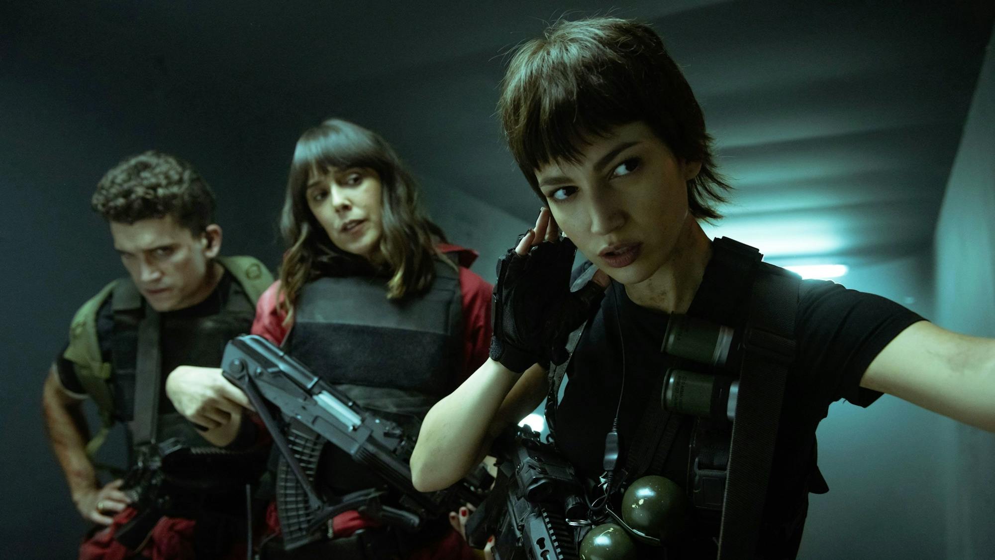Denver (Jaime Lorente), Malina (Belén Cuesta), and Tokio (Úrsula Corberó) stand in a poorly lit hallway wearing bullet proof vests and carrying guns and grenades.