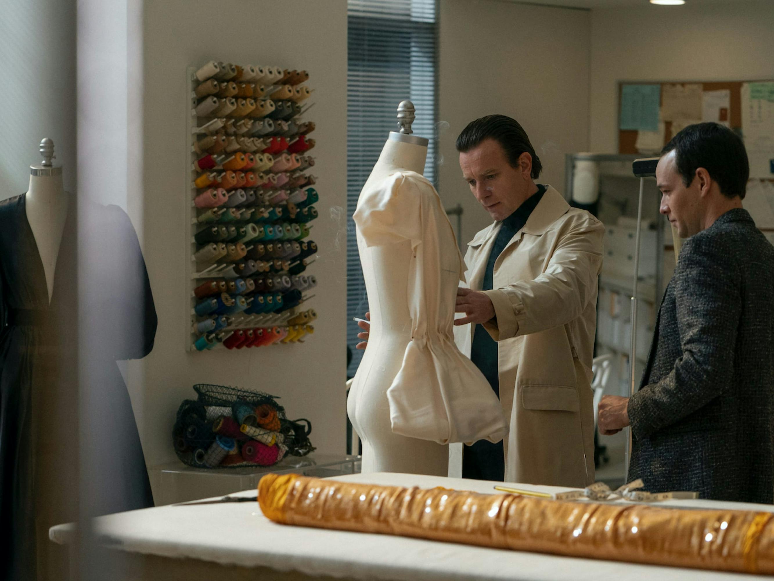 Halston (McGregor) stands in his studio with another person tending to a mannequin. He wears a tan coat and black turtleneck. His companion is in a plaid blazer. There are spools of thread behind them, and the mannequin in question appears to wear a white shirt.