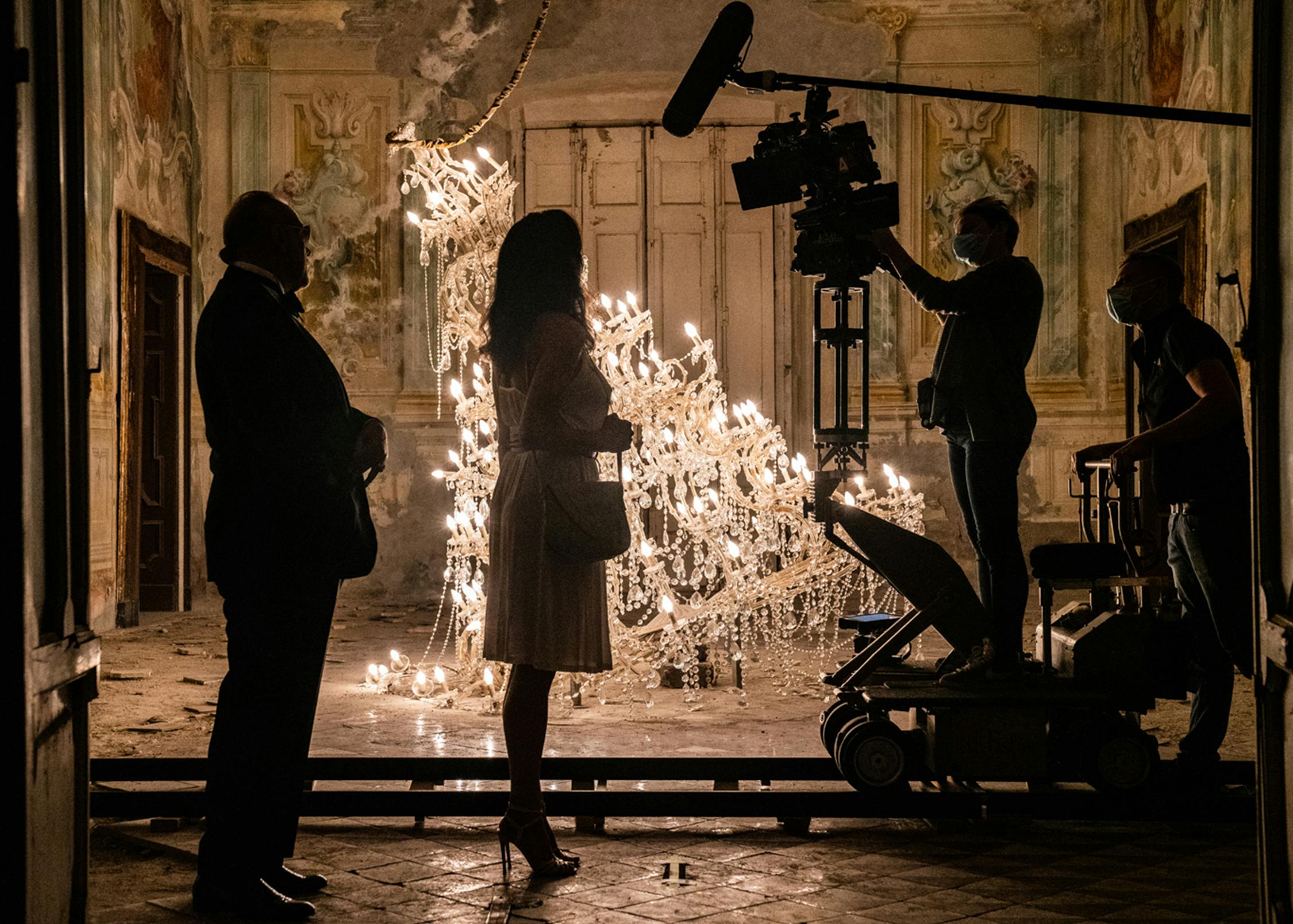 The shadowy figures of the cast and crew of The Hand of God stand beside a fallen chandelier. On the walls is ornate antique molding. 