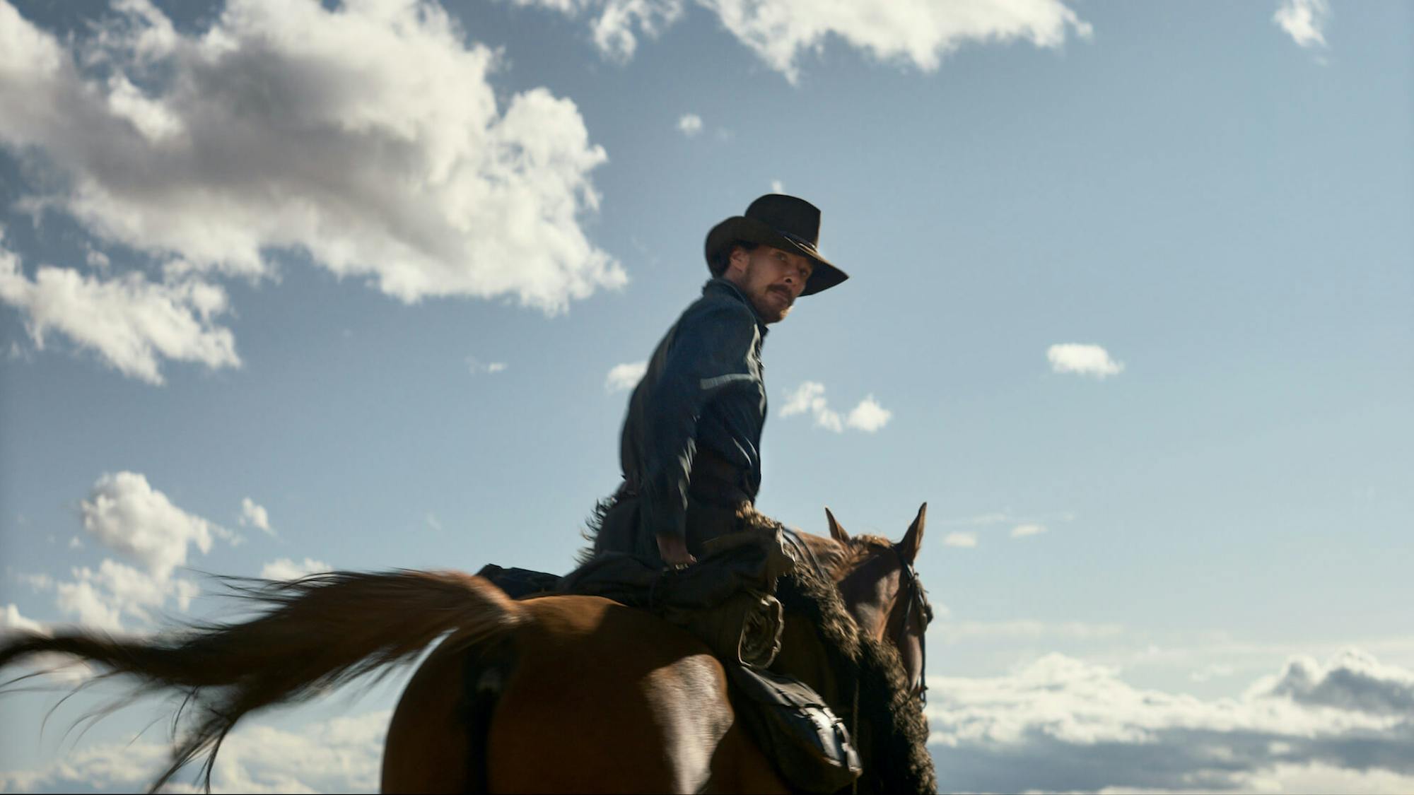 Phil Burbank (Benedict Cumberbatch) rides a brown horse in a denim shirt and brown hat. The sky is bright blue and streaked with puffy clouds.