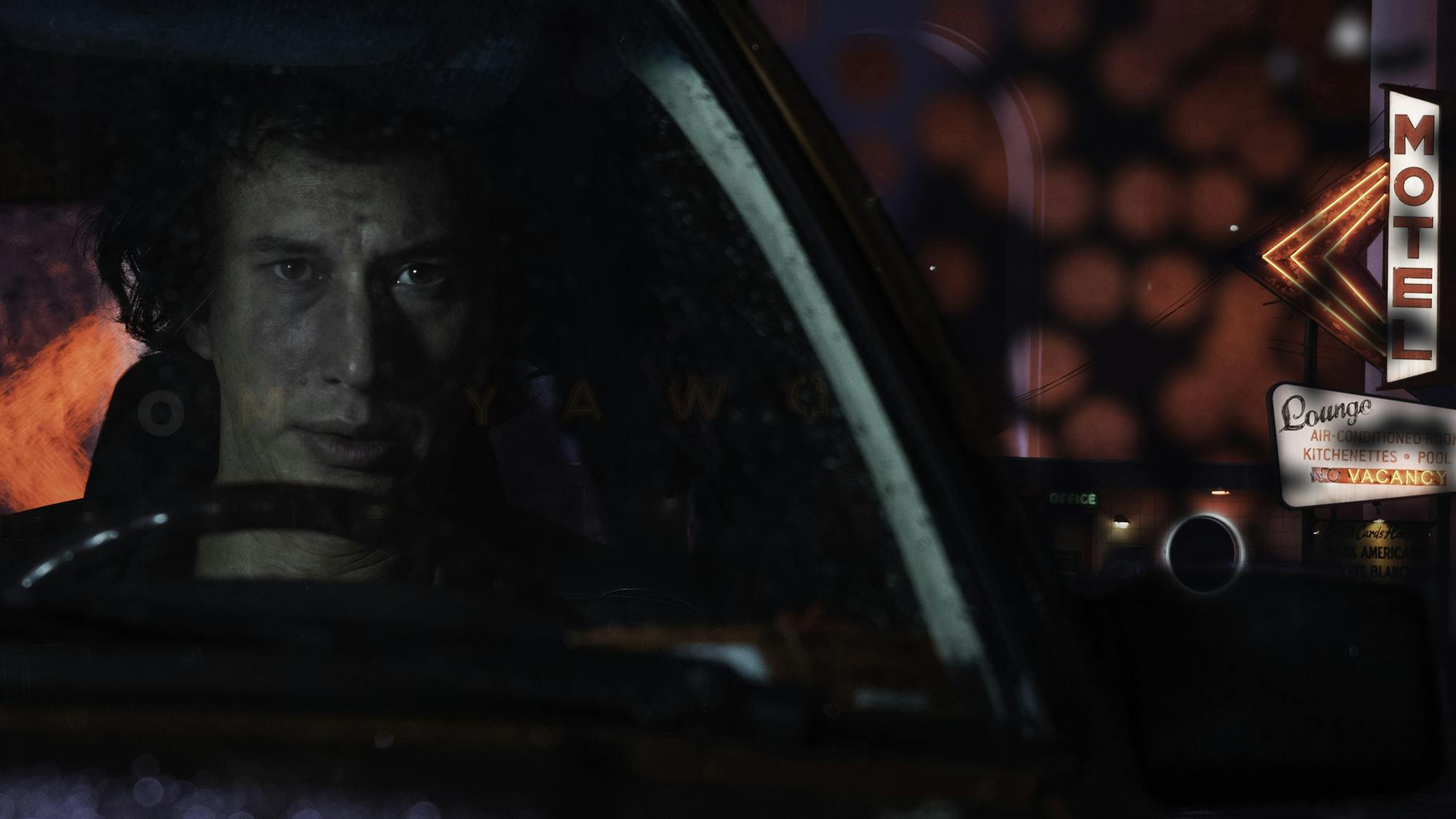 Jack Gladney (Adam Driver) sits in a car at night outside of a motel, there is rain on the windshield and a red glow from neon lights behind his head as he stares intensely forwards. 