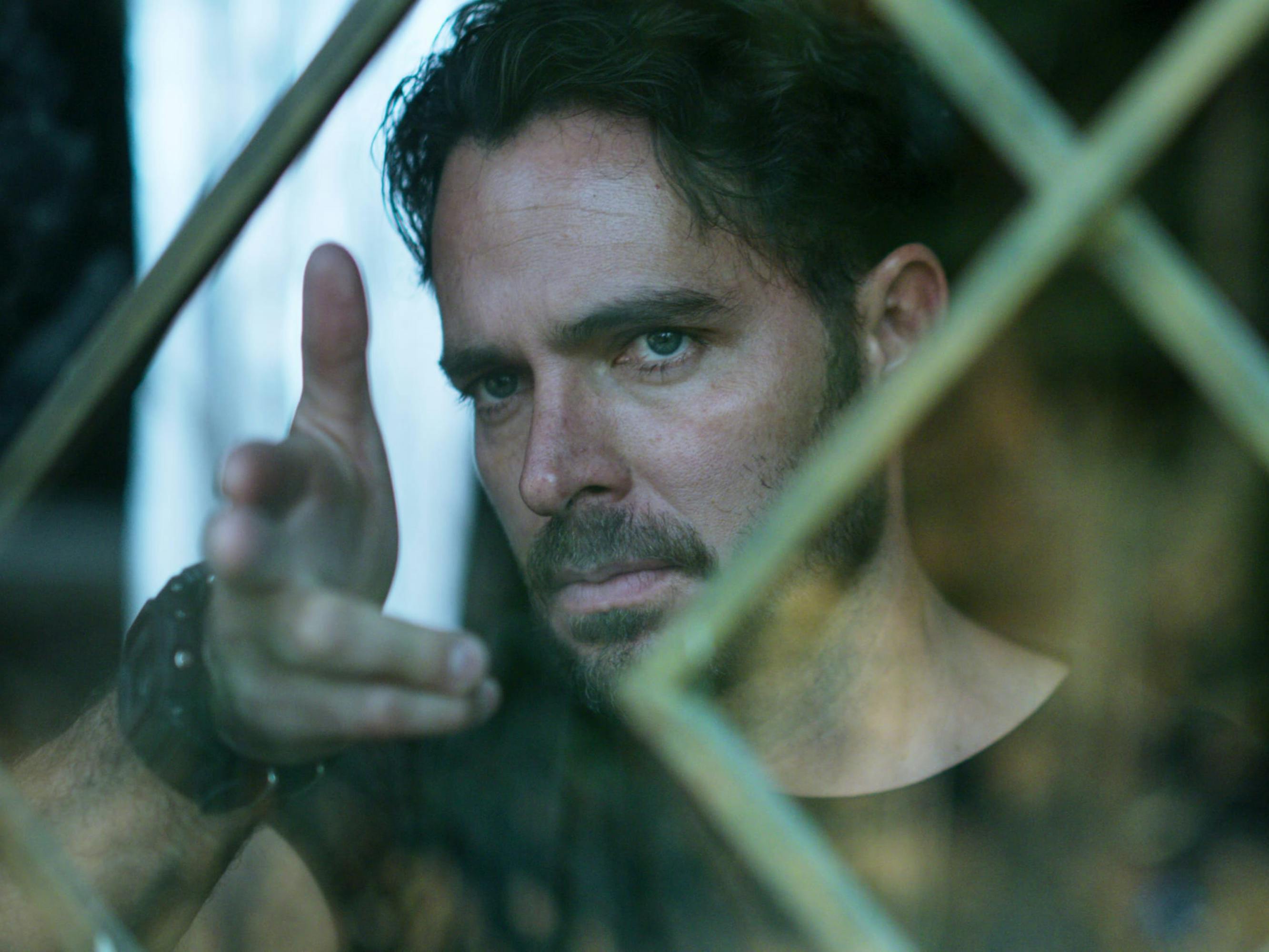 Actor Manolo Cardona who plays Sara’s brother, Álex, stands behind a barred window. He wears a black t-shirt and a chunky black watch. He also sports some facial hair. His fingers are poised in the shape of a gun, and he does not look happy.