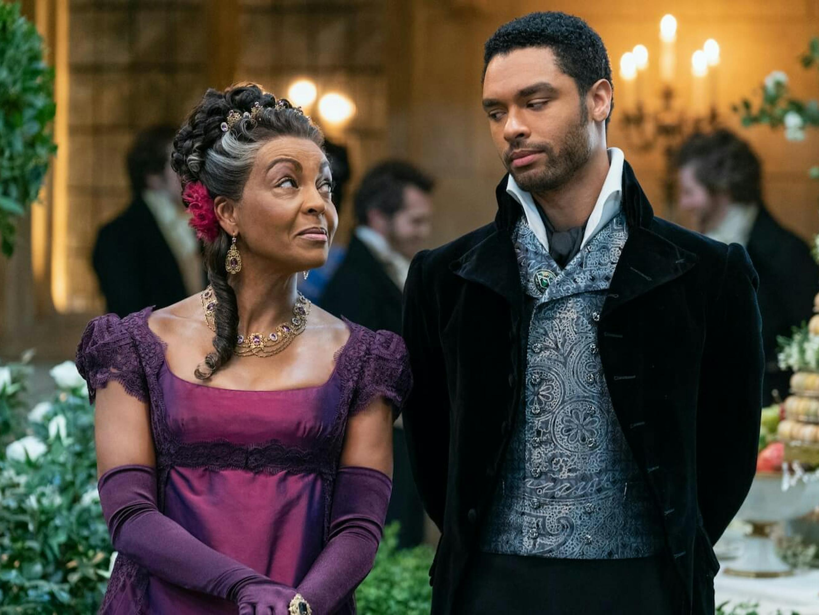 Lady Danbury (Adjoa Andoh) and Simon Basset (Regé-Jean Page) walk through a ballroom scene, filled with lights, flowers, and little treats. Lady Danbury looks teasingly at her pseudo-son, and Basset looks back. Basset looks studly as ever in a black velvet blazer, paisley jacket, and high white collar. Danbury wears a shimmery purple and pink dress, with a pink flower behind her ear.