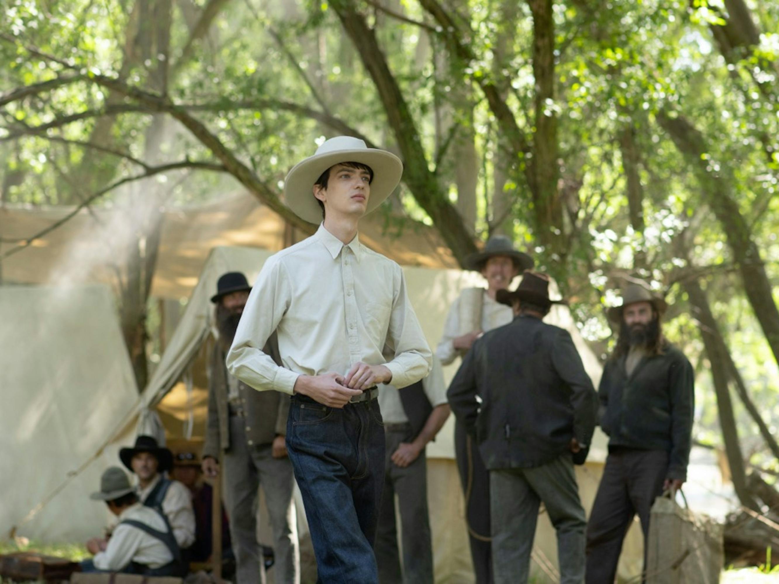 Kodi Smit-McPhee’s character Peter walks around a campsite in a white hat, white shirt, and dark pants. In the background mingle men in dark jackets, hats, and white shirts. Dappled sunlight lits this Western scene.
