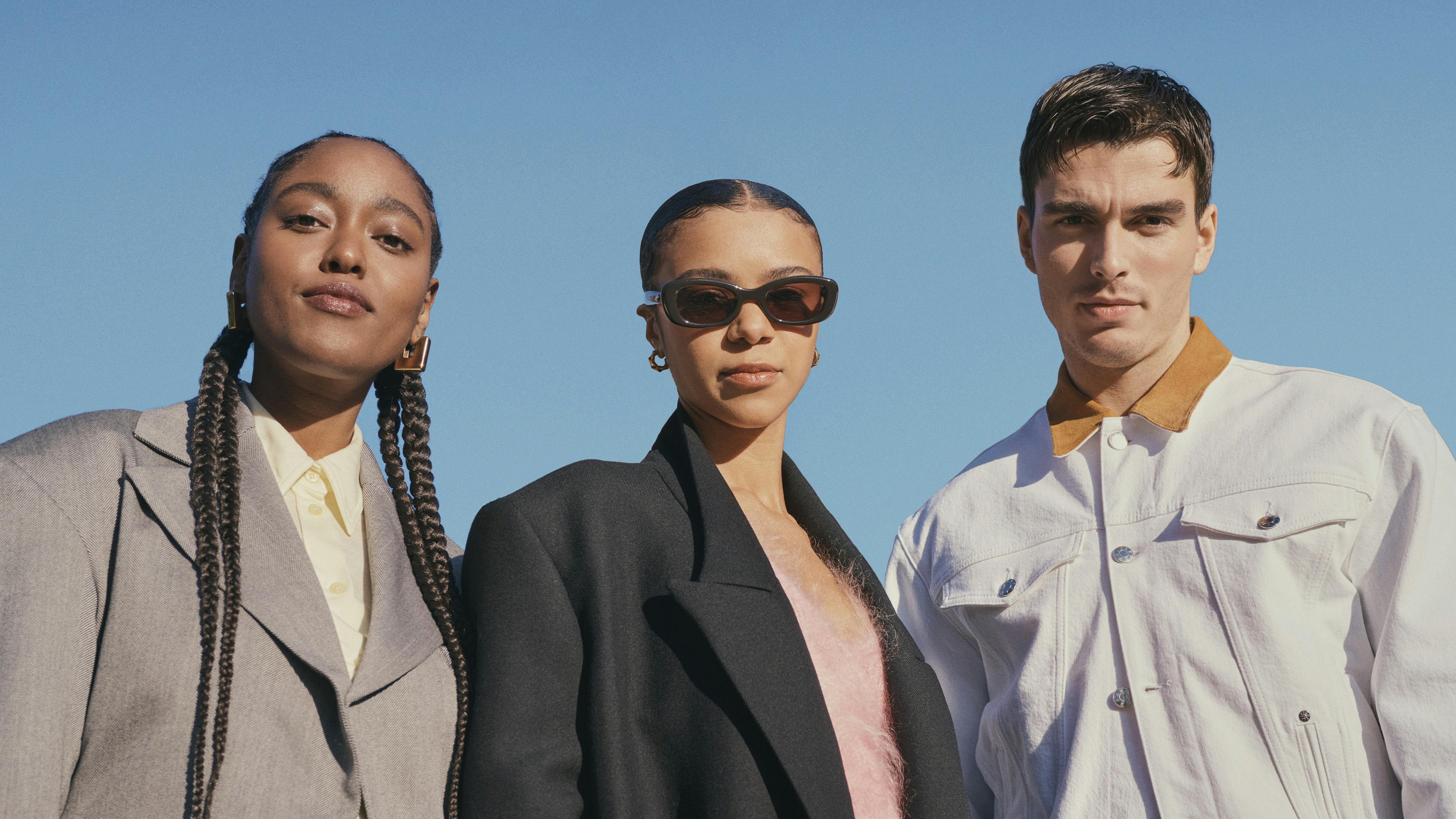 Arsema Thomas, India Amarteifio, and Corey Mylchreest stand together against a blue sky. Thomas wears jeans and a gray blazer. Amarteifio wears a black jacket and sunglasses. Mylchreest wears a white jacket and black pants.