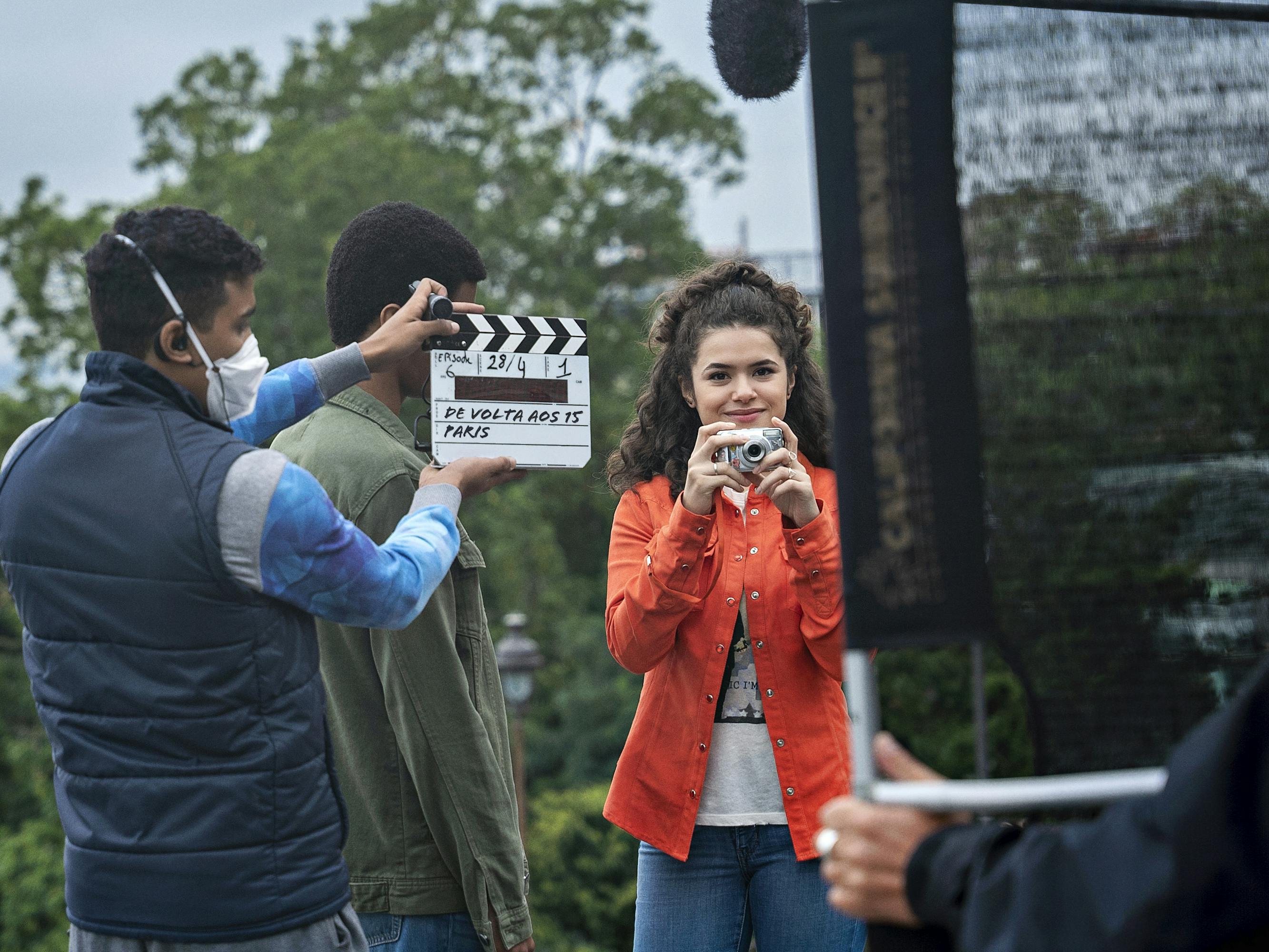 Maisa Silva wears jeans, a white t-shirt, and an orange jacket. She mingles with crew members, one of whom holds a clapperboard.