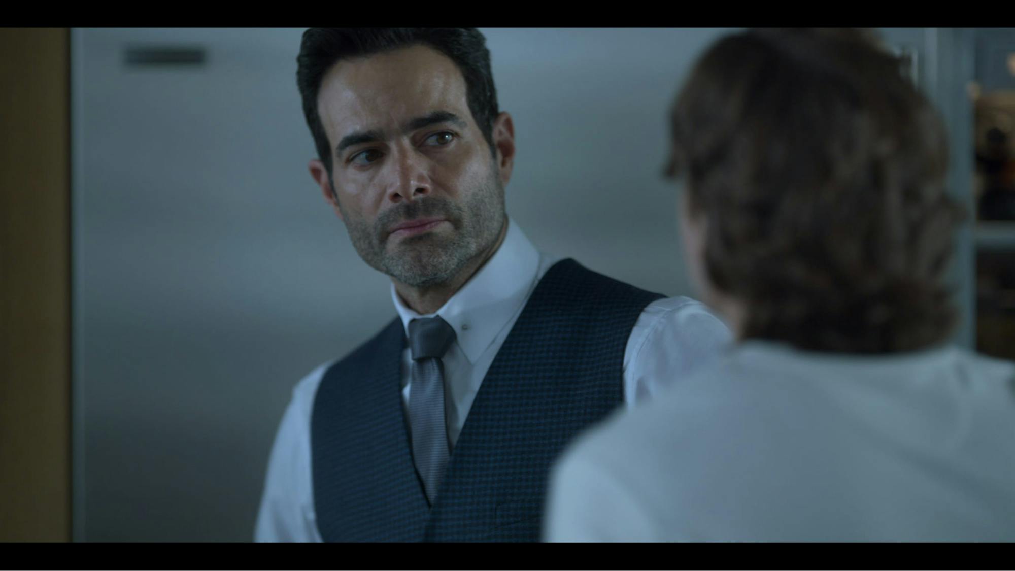 Lorenzo (Luis Roberto Guzmán) wears a white shirt, grey tie, and navy vest. He looks scared as he faces a man in the foreground of this shot.