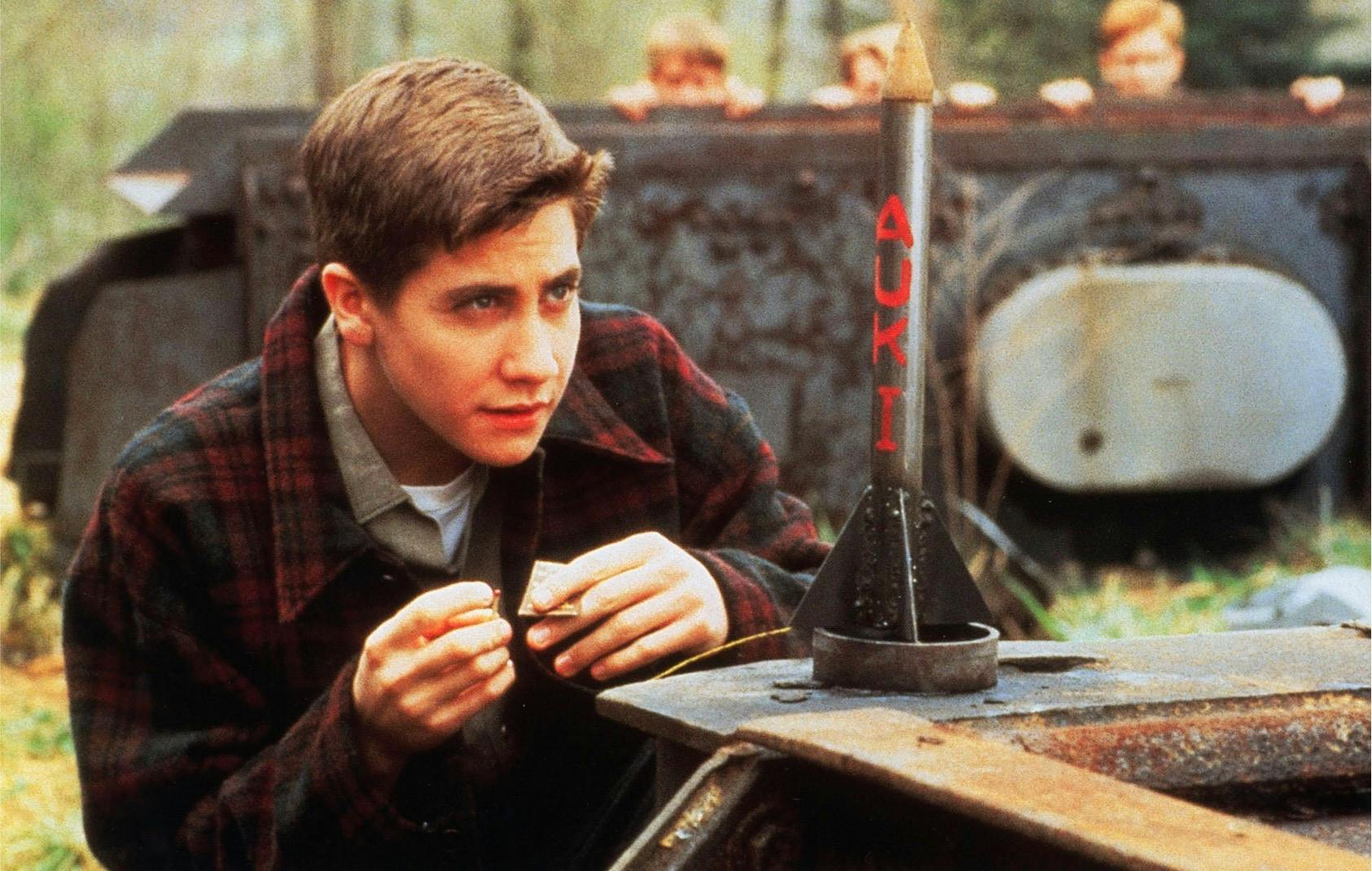 Homer Hickman (Jake Gyllenhaal) in October Sky (1999) crouches beside a homemade rocket. He wears a plaid flannel jacket and looks excited and focused.