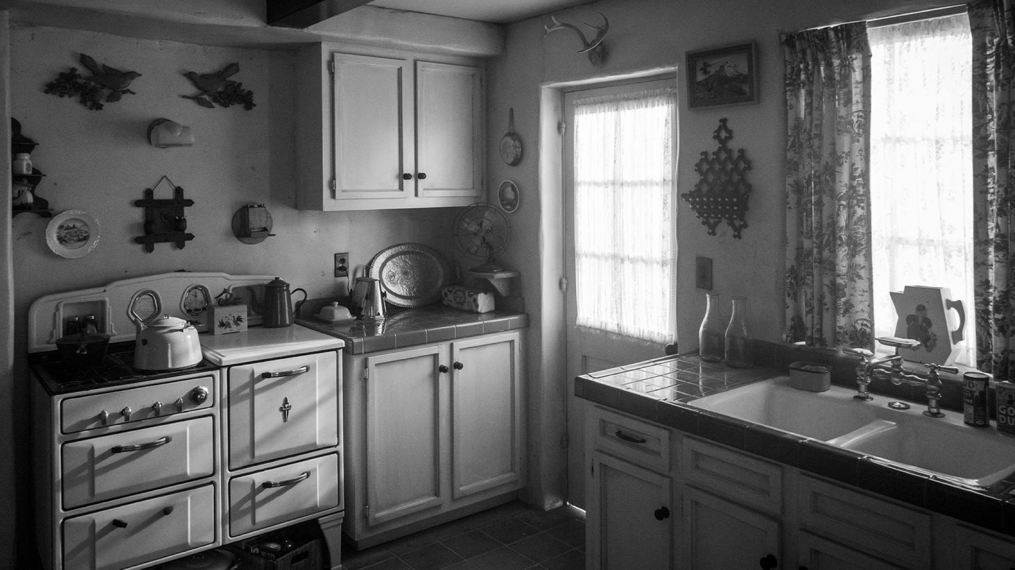 The kitchen set at the Victorville Bungalow. The room is small. The floor and counters are tiled. The walls are decorated with a quaint assortment: a small landscape painting, a decorative plate, a diminutive set of antlers. Light floods in from outside, filtered through sheer curtains. 