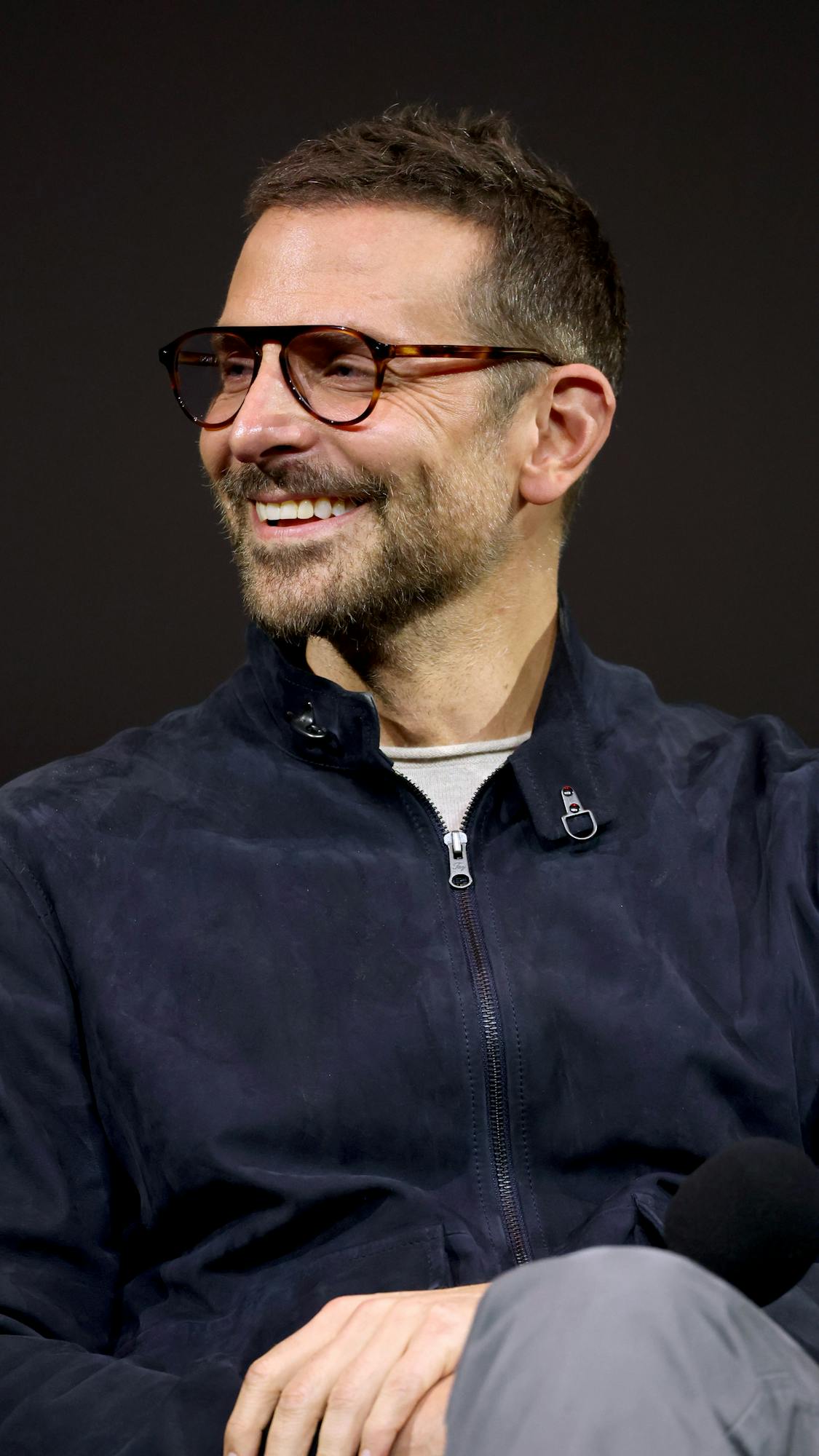 Bradley Cooper wears a navy jacket and brown glasses.