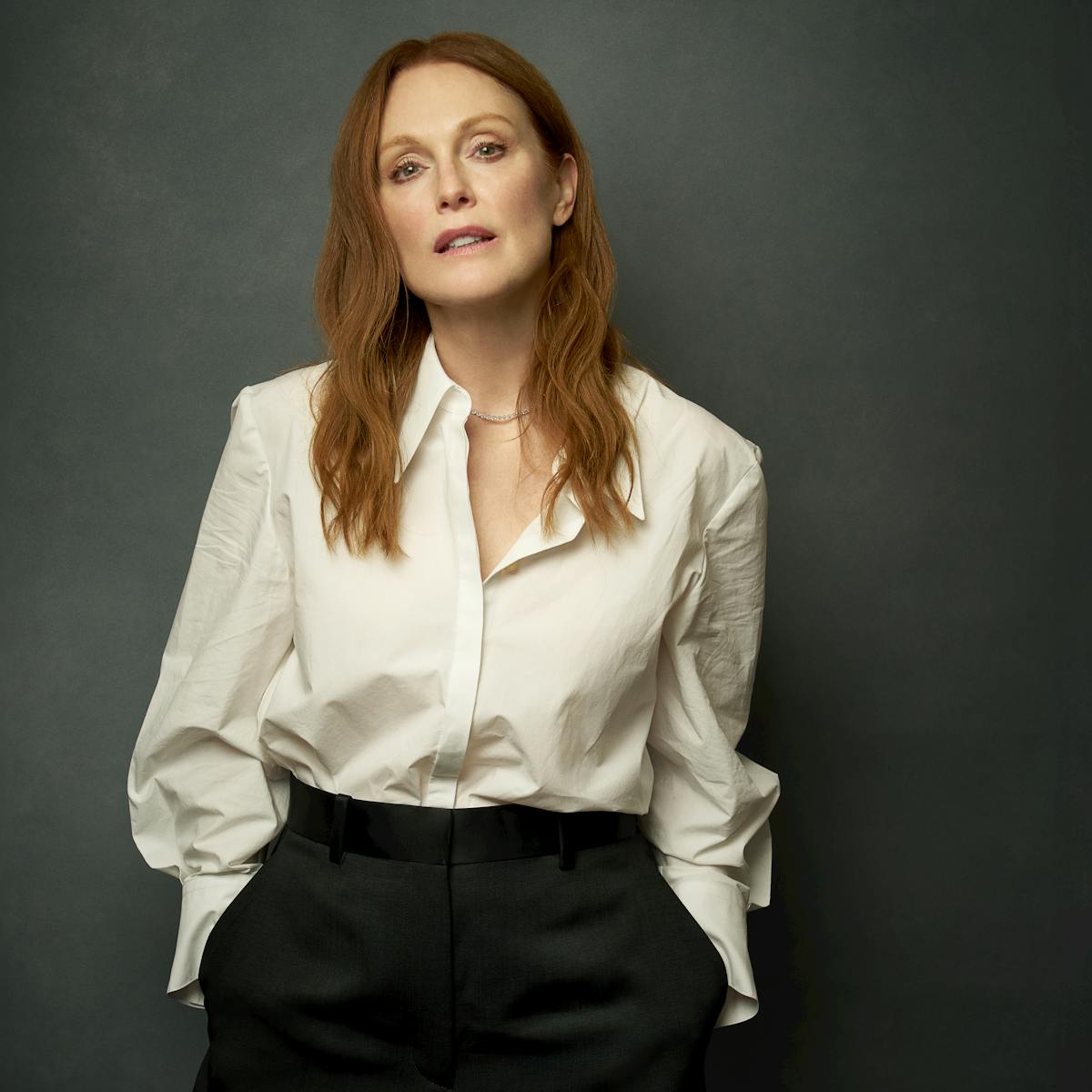 Julianne Moore wears a white shirt and black pants and stands in front of a dark background. 