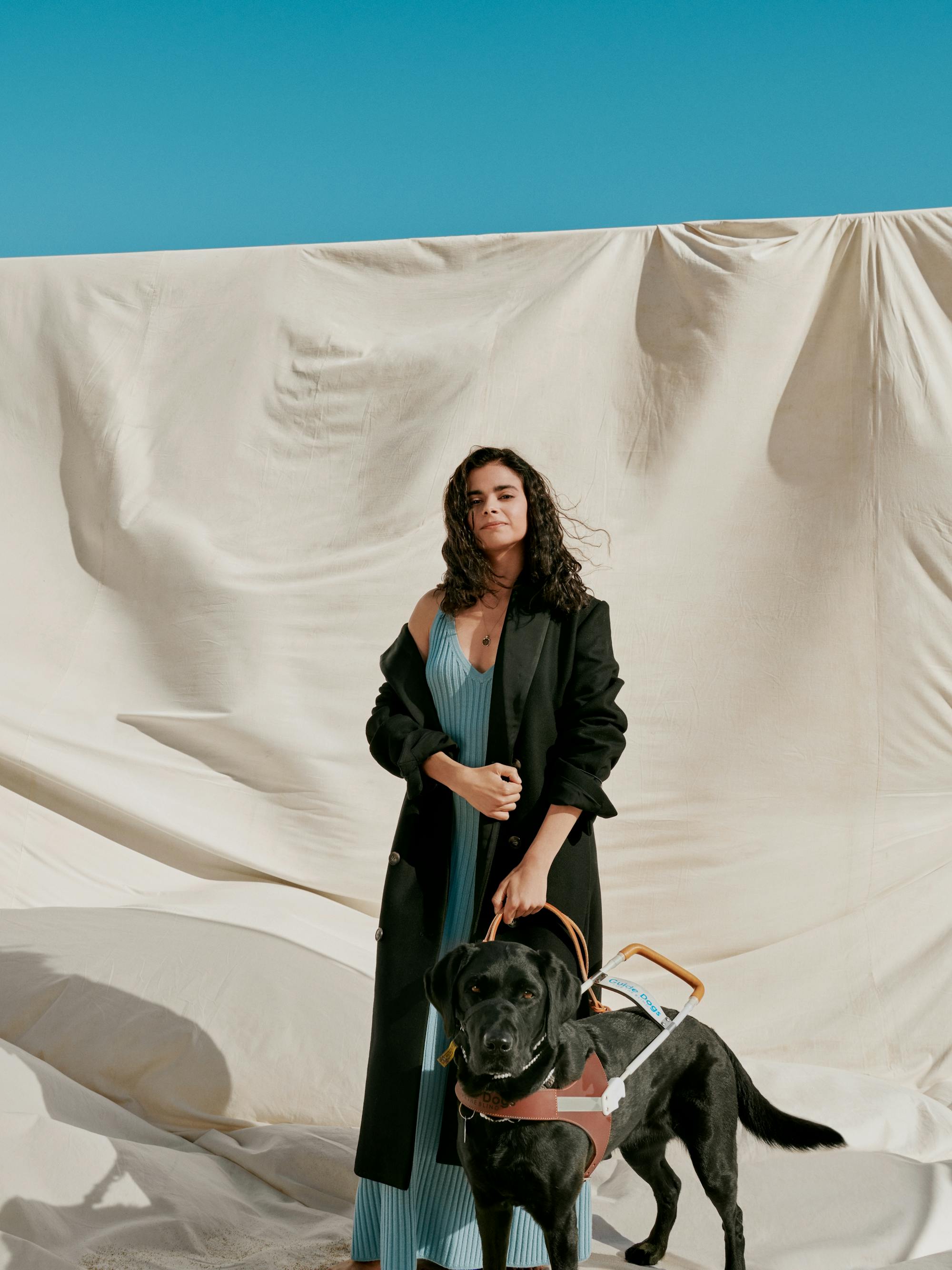 Aria Mia Loberti wears a blue dress and long black jacket, and holds the harness of her black lab.