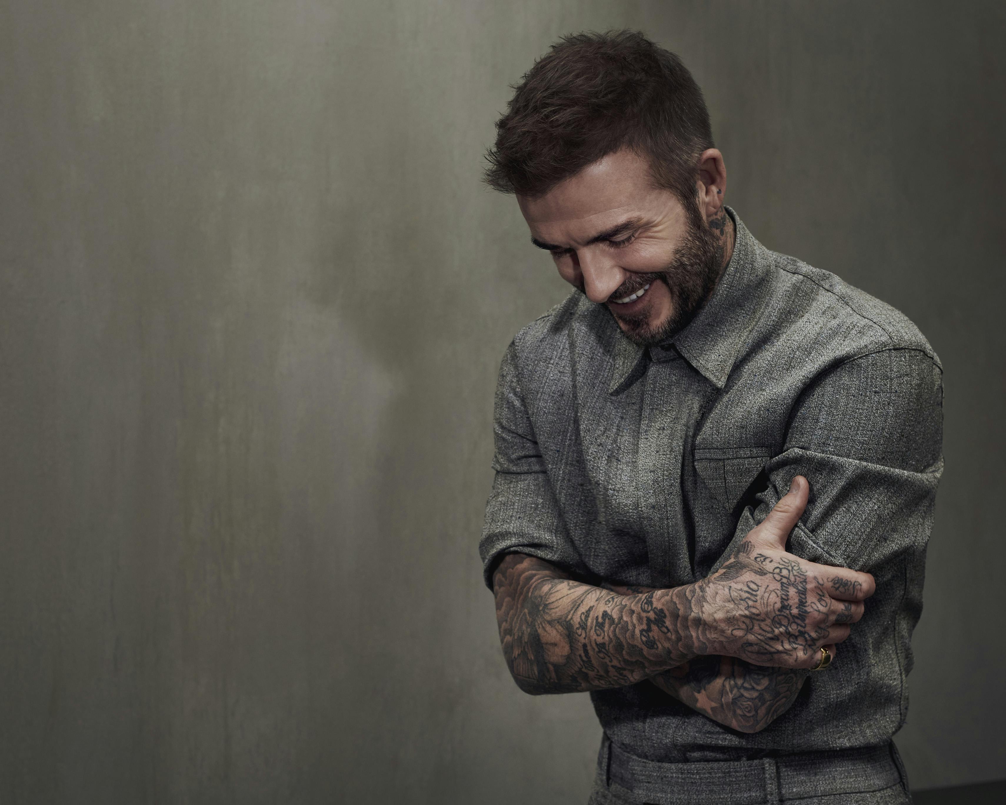 David Beckham wears a gray shirt and pants and shows off his tattoo sleeve against a gray wall. 