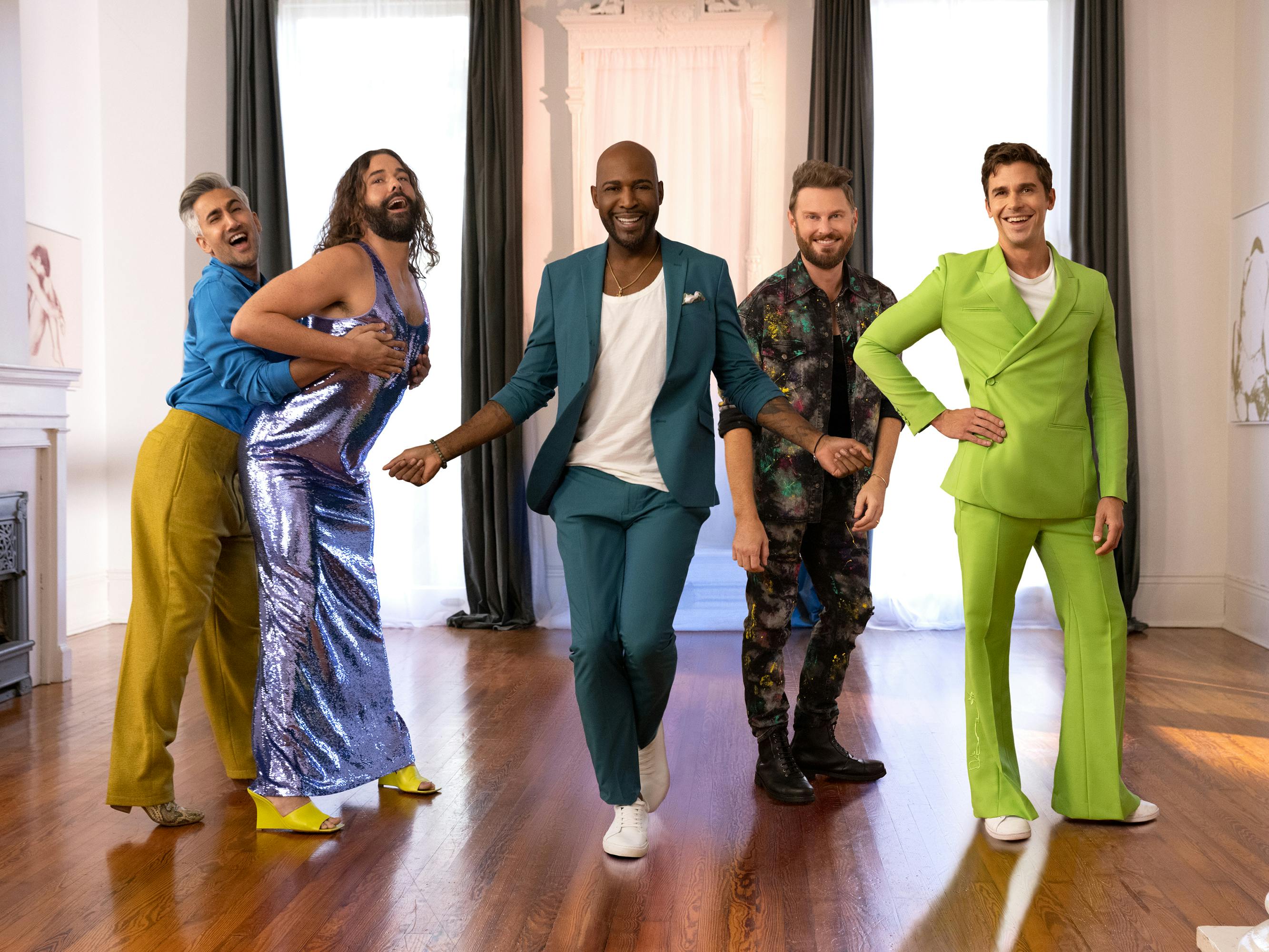 The Fab Five — Tan France, Jonathan Van Ness, Karamo, Bobby Berk, and Antoni Porowski. Each wears a bright outfit: Tan pairs a bright blue shirt with yellow pants, Van Ness glitters in a jumpsuit and yellow shoes, Karamo strikes a pose in a teal suit, Berk wears a matching set in a chaotic pattern, and Porowski wears an extremely tailored lime green double-breasted suit.