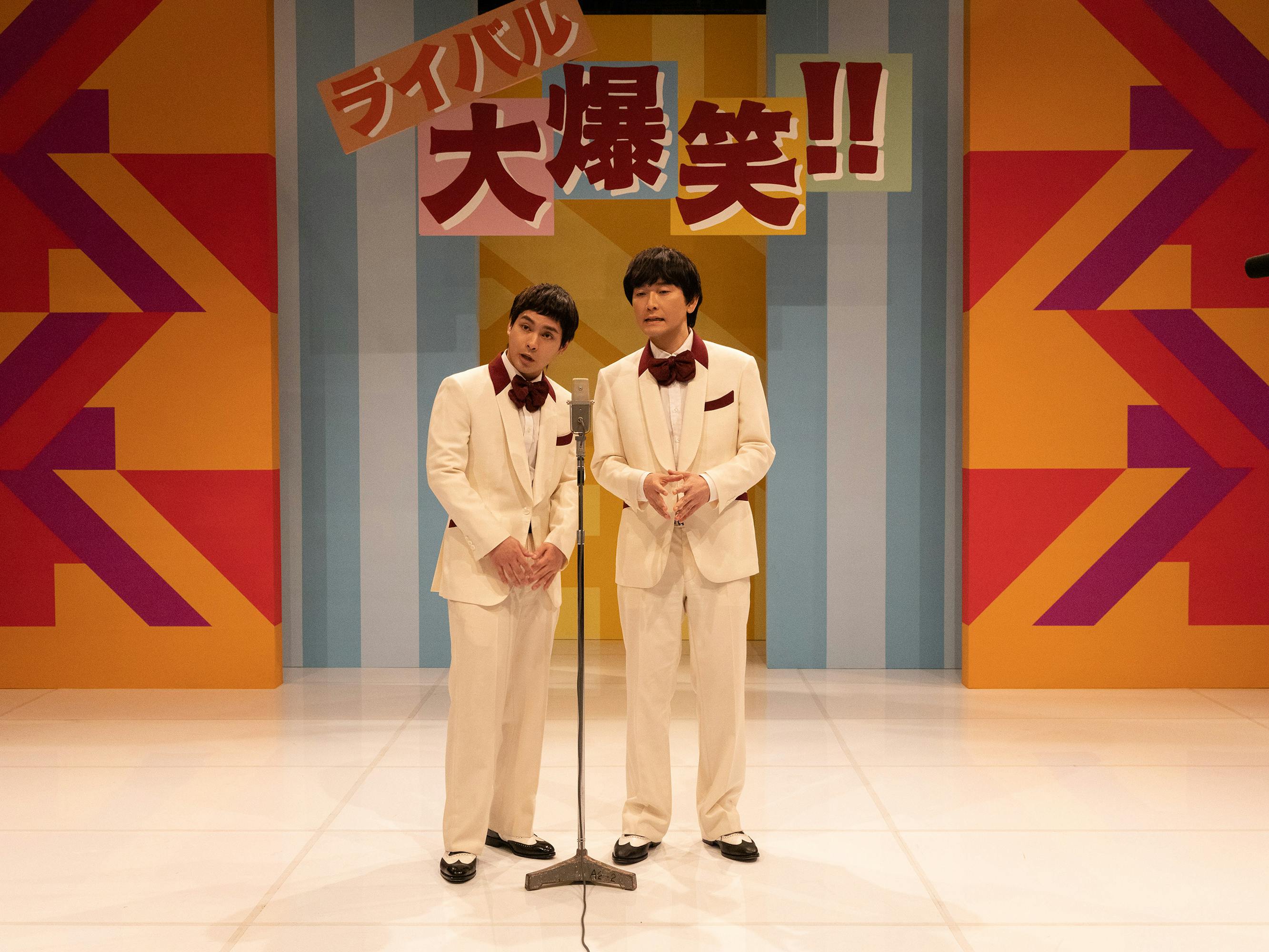 Yuya Yagira and Nobuyuki Tsuchiya wear white suits on stage. Behind them the walls are orange, red, and blue. 