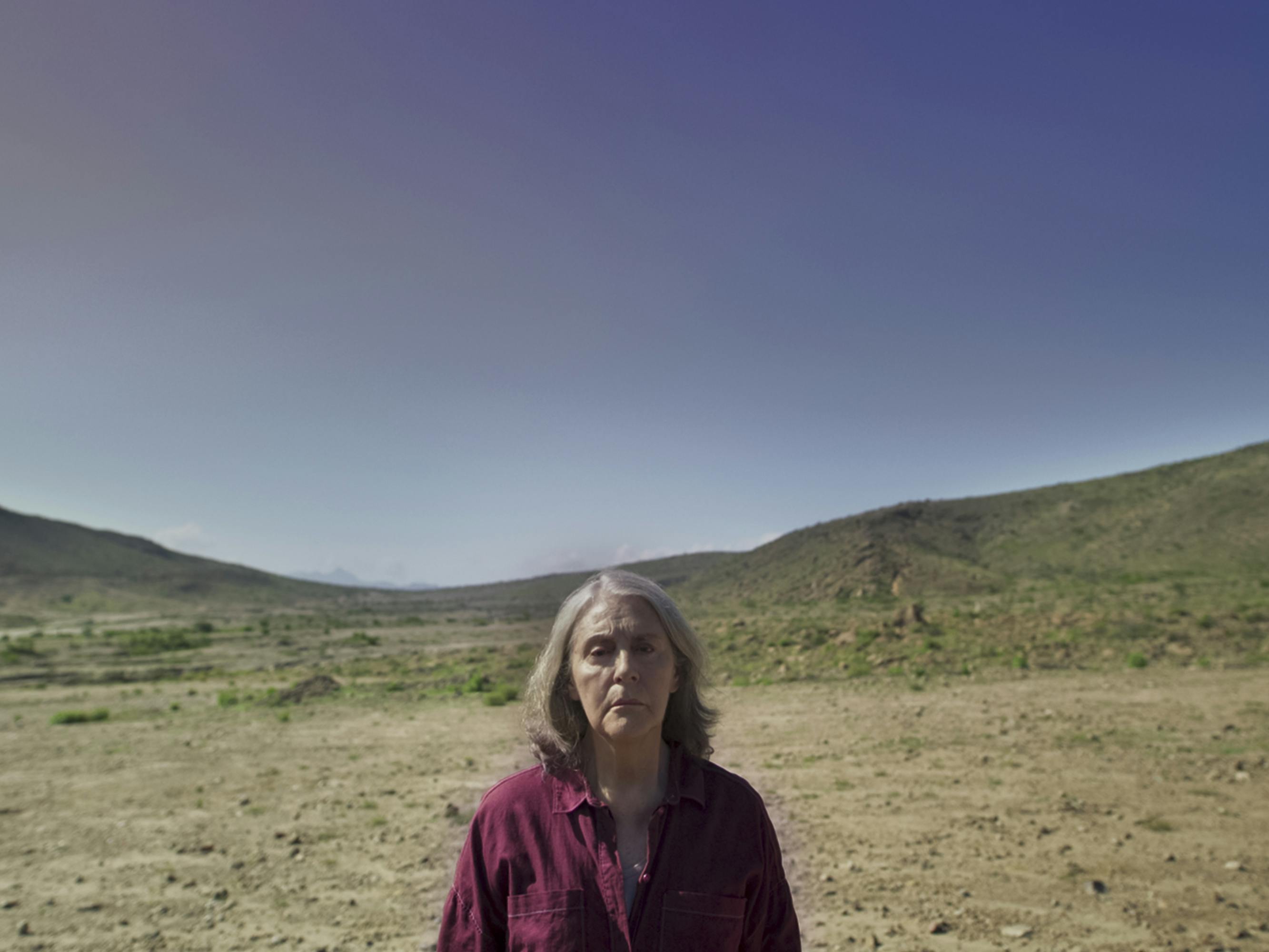 A woman wears a red shirt and stands in a barren landscape against a bright blue sky.