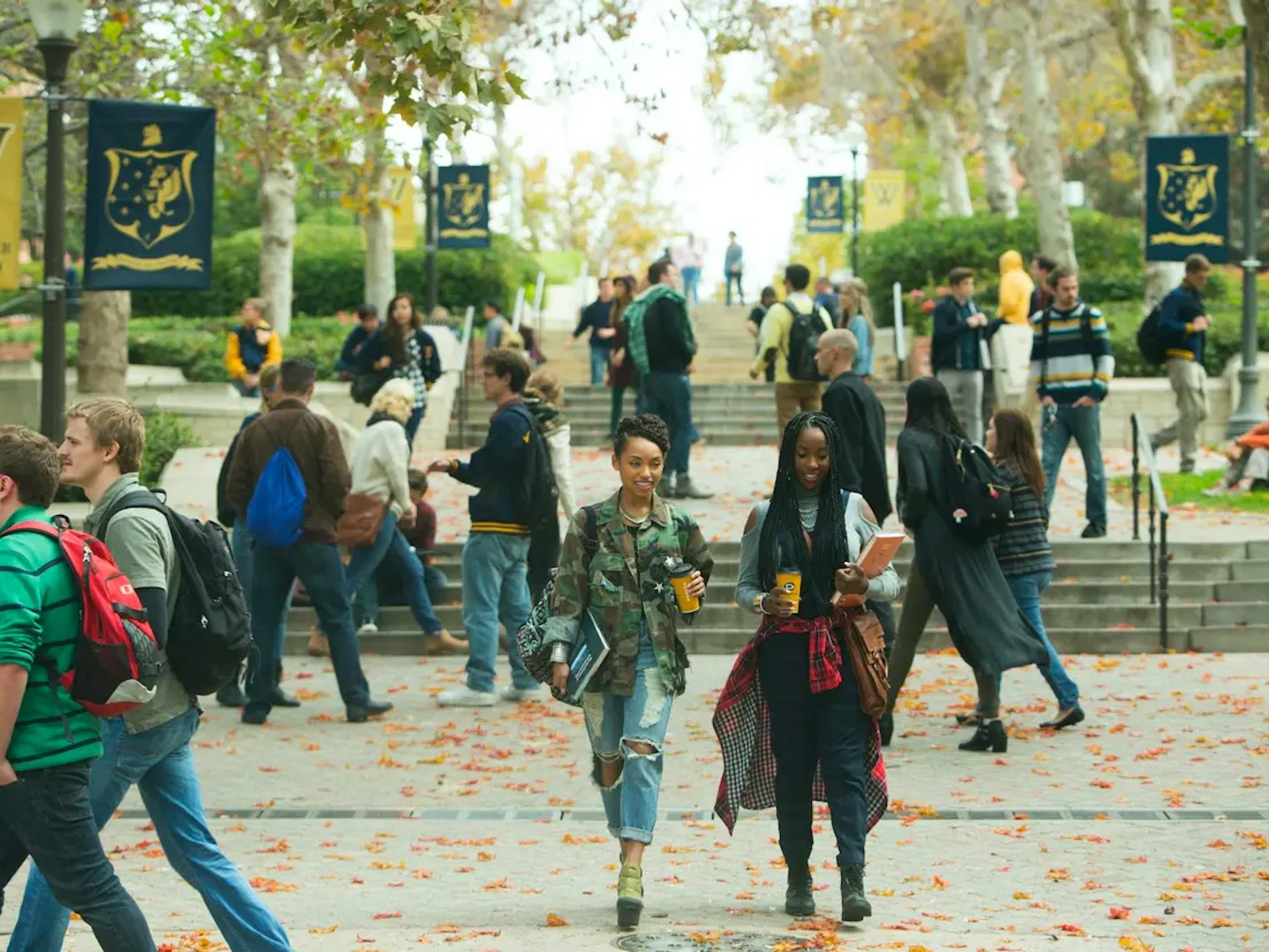 Sam White (Logan Browning) and Joelle Brooks (Ashley Blaine Featherson) walk through a collegial scene. There are leaves on the ground and students walking through. The two hold coffee and carry books.