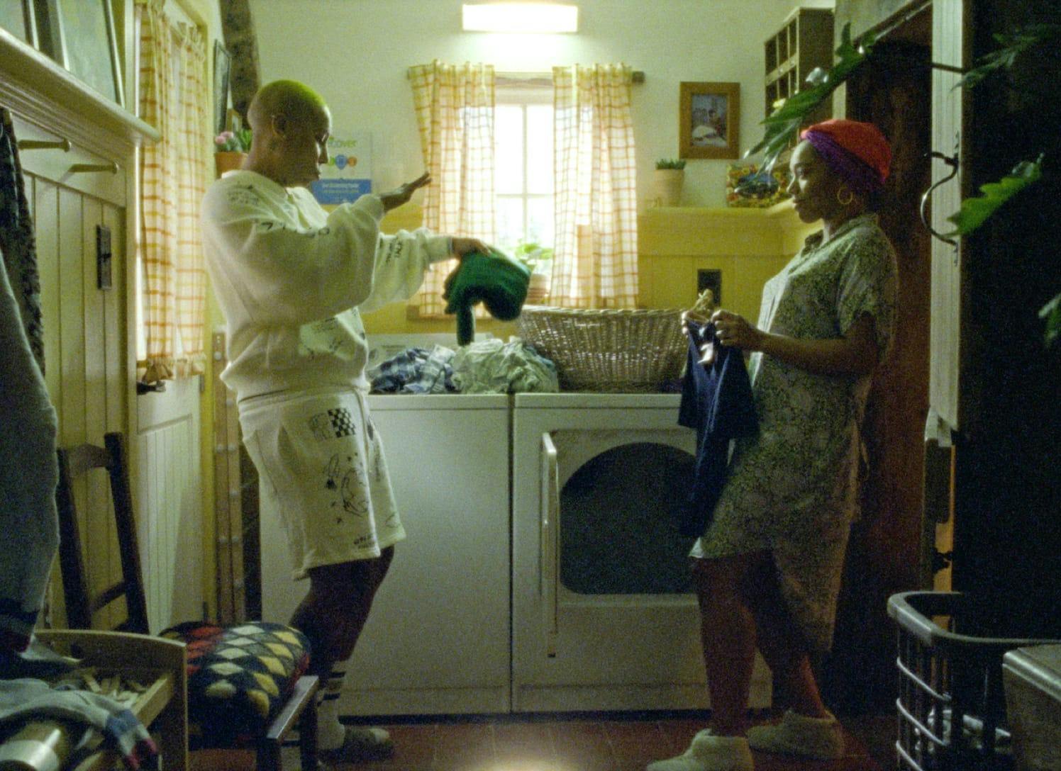 Denise (Lena Waithe) and Alicia (Naomi Ackie) dance while doing laundry. Denise holds a green garment, and Alicia holds a blue one. There is a basket and pile of clothes atop the machines.