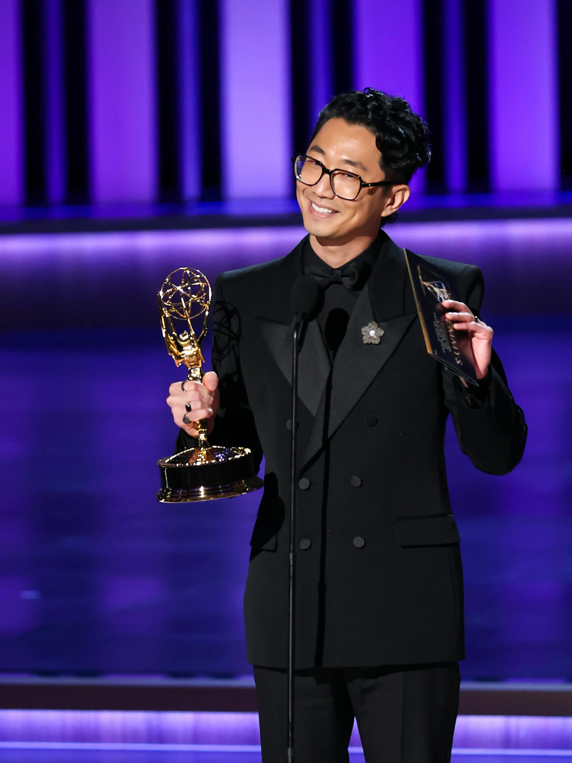 Lee Sung Jin wears all black and holds his Emmy on a purple stage.
