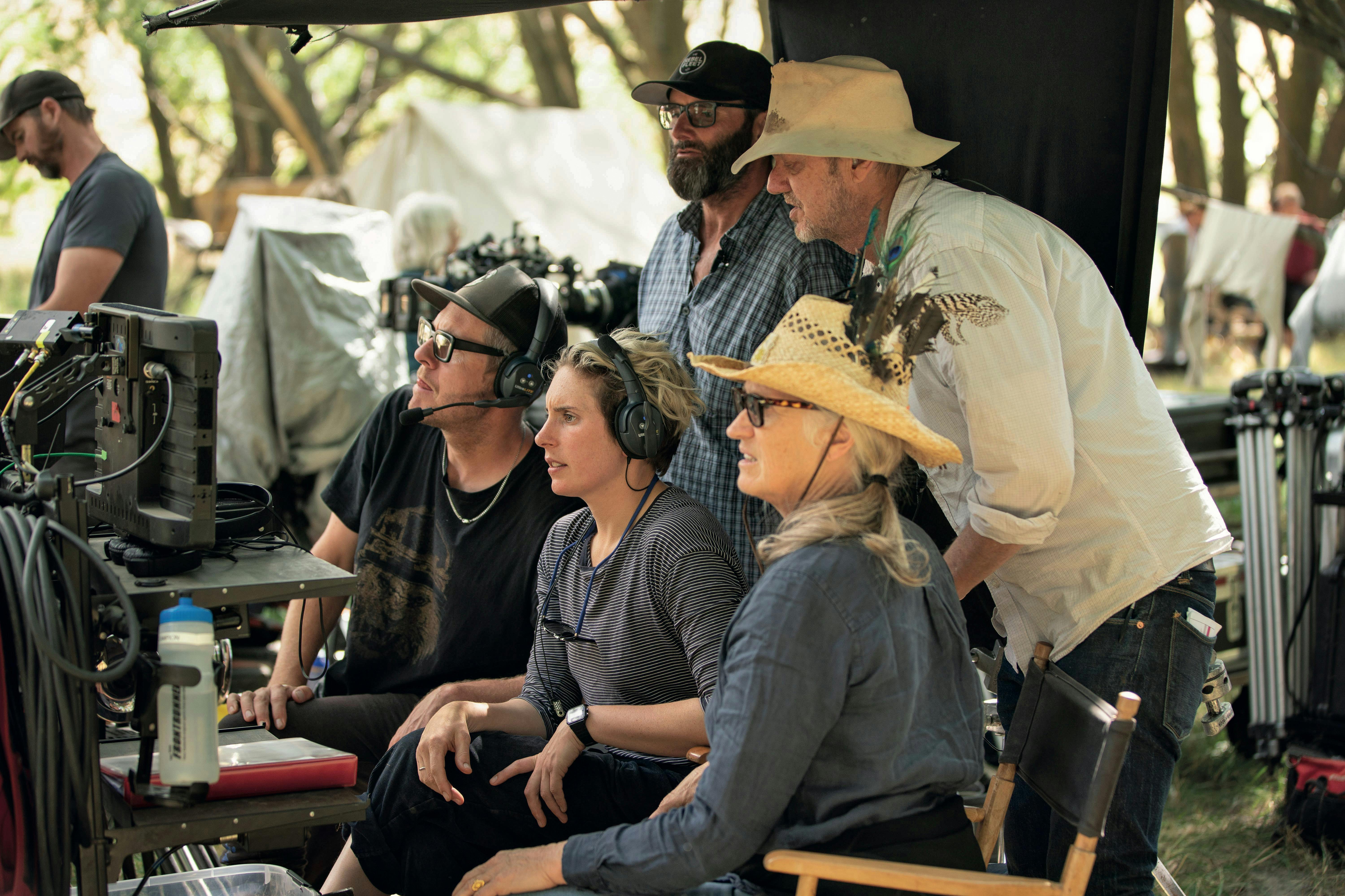 Ari Wegner, Jane Campion, and The Power of the Dog crew sit behind a camera rig. The team wears an assortment of straw hats and headphones, with Jane's feathered cap as a standout.