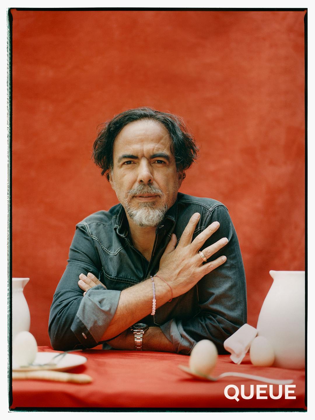 Alejandro González Iñárritu wears a denim shirt and sits against a red drape. In front of him are white ceramics and some eggs.