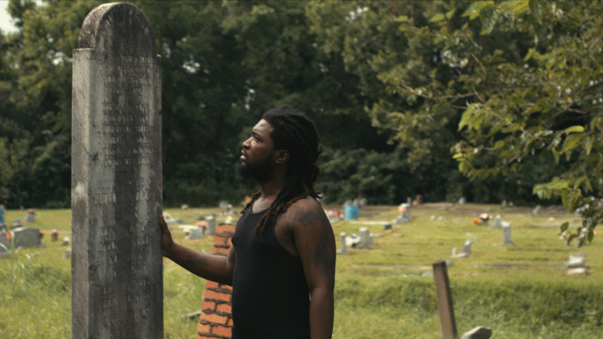 Emmett Lewis wears a black tank top and stands next to a worn-down gravestone in a grassy graveyard.