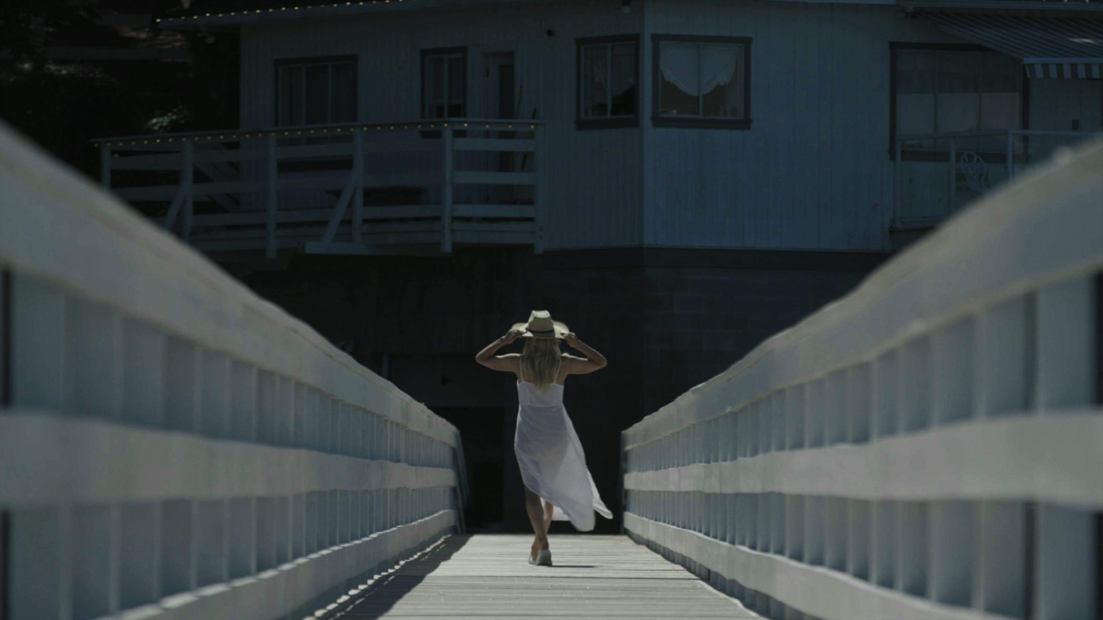 Pamela Anderson wears a white dress and straw hat and walks down a dock.