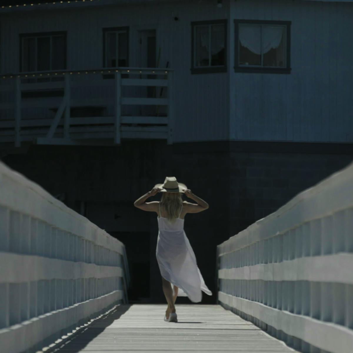 Pamela Anderson wears a white dress and straw hat and walks down a dock.
