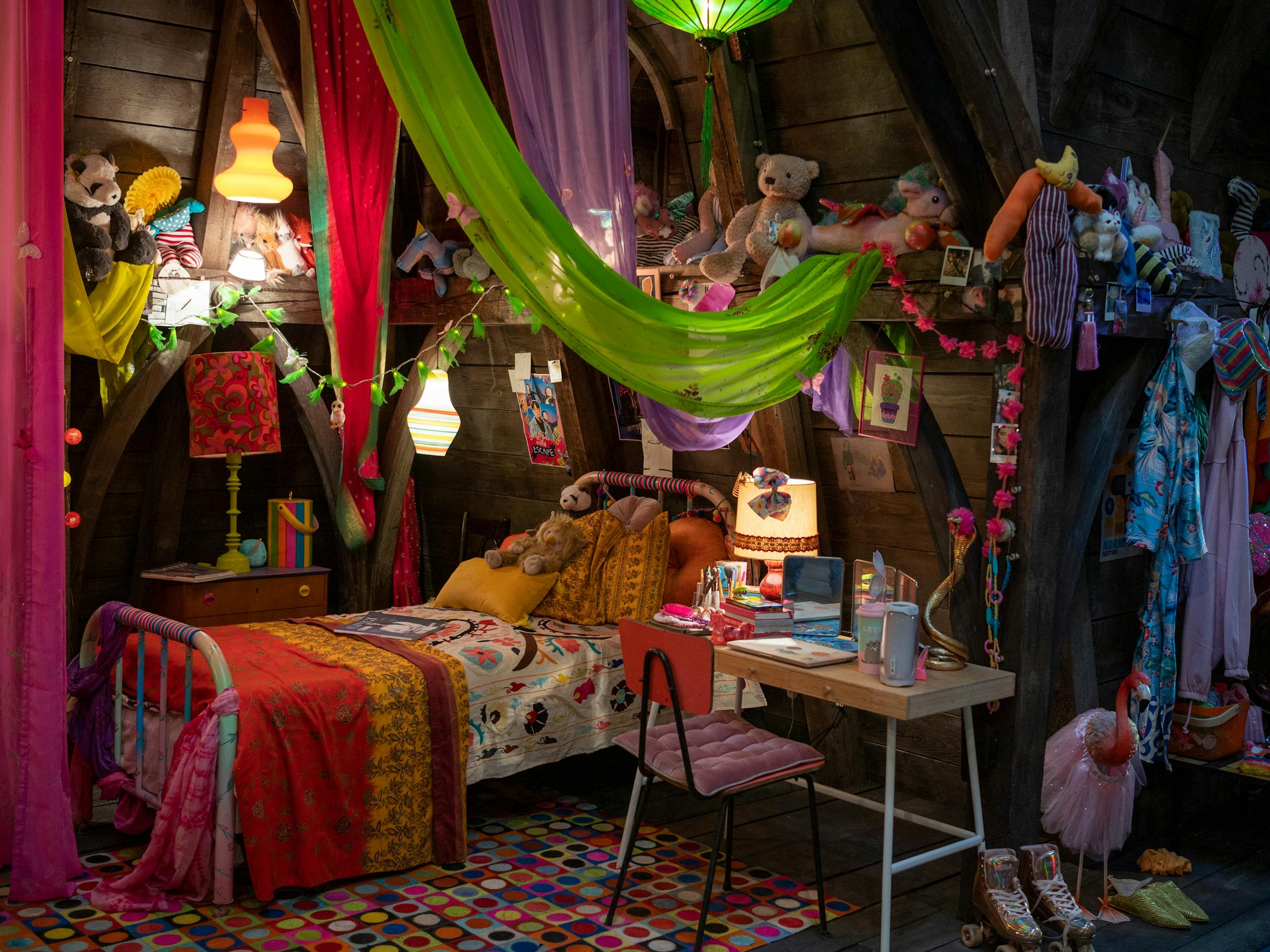Enid Sinclair's (Emma Myers) side of the room. There is a multicolored rug, a green-pink-and-purple awning, lots of colorful clothes, trinkets and knick knacks, and a colorful quilt atop a cozy-looking bed.