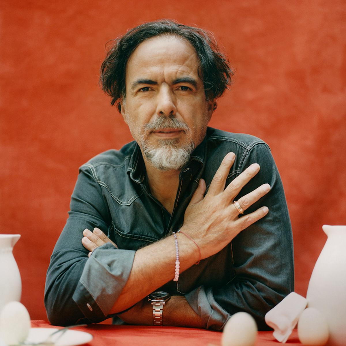 Alejandro González Iñárritu wears a denim shirt and sits against a red drape. In front of him are white ceramics and some eggs.