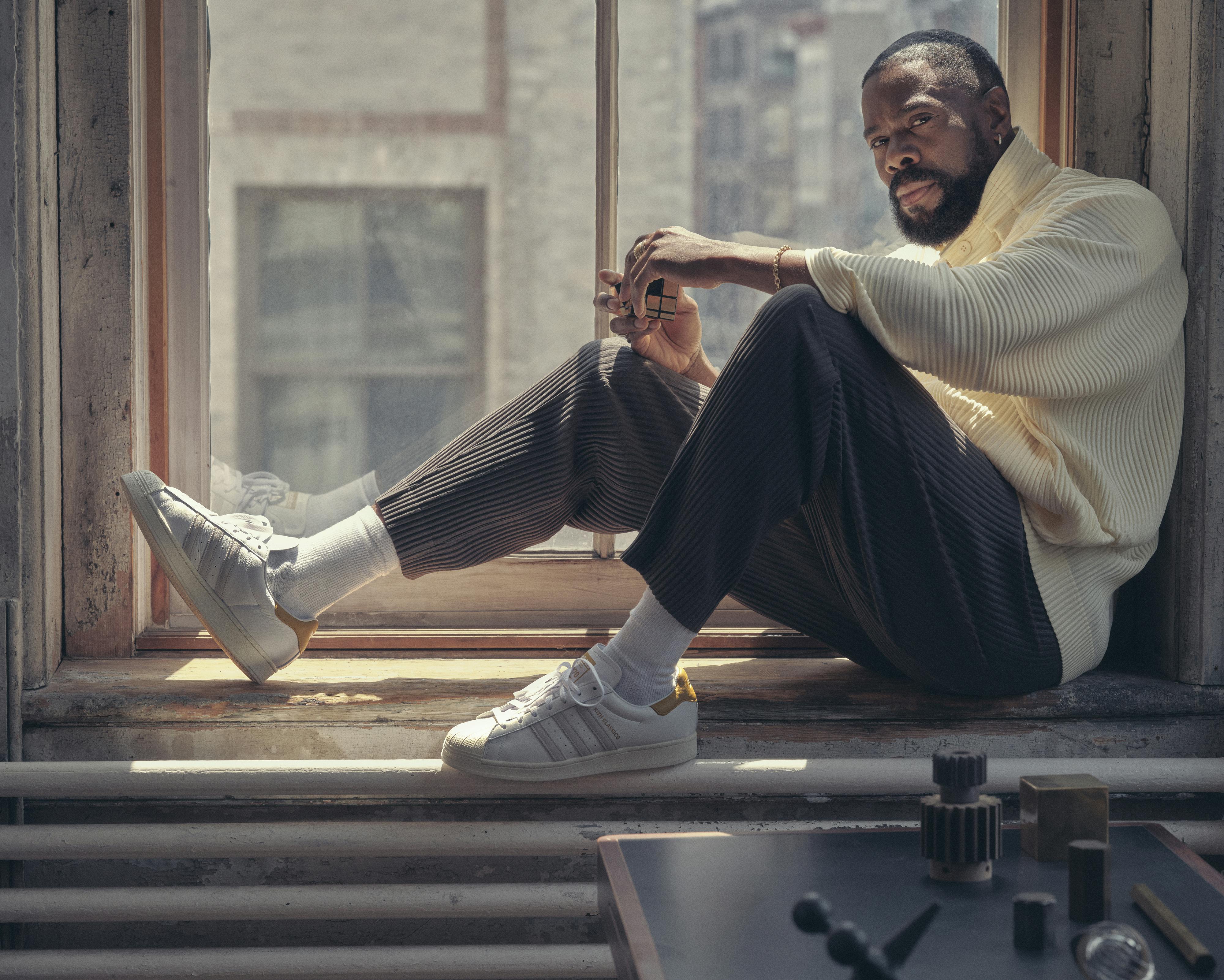Colman Domingo wears dark pants, a cream sweater, and white sneakers as he sits on a windowsill.
