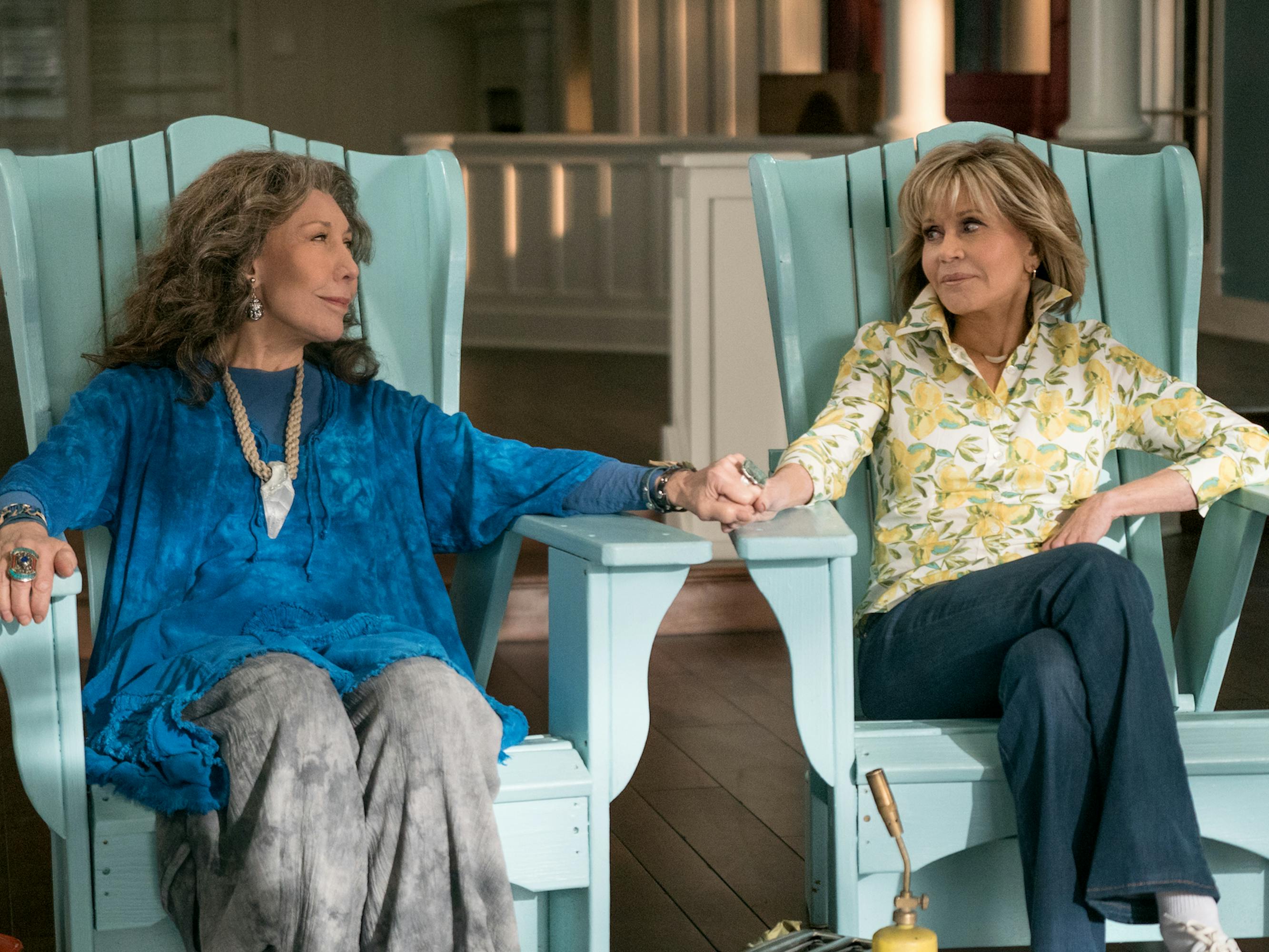 Frankie (Lily Tomlin) and Grace (Jane Fonda) sit in turquoise chairs and hold hands across the arm rests.