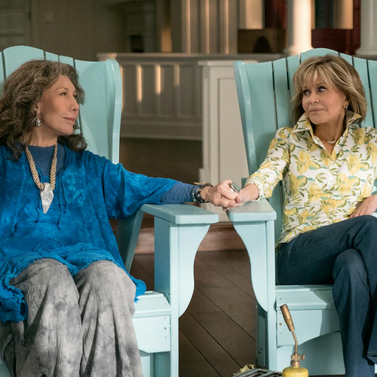 Frankie (Lily Tomlin) and Grace (Jane Fonda) sit in turquoise chairs holding hands.