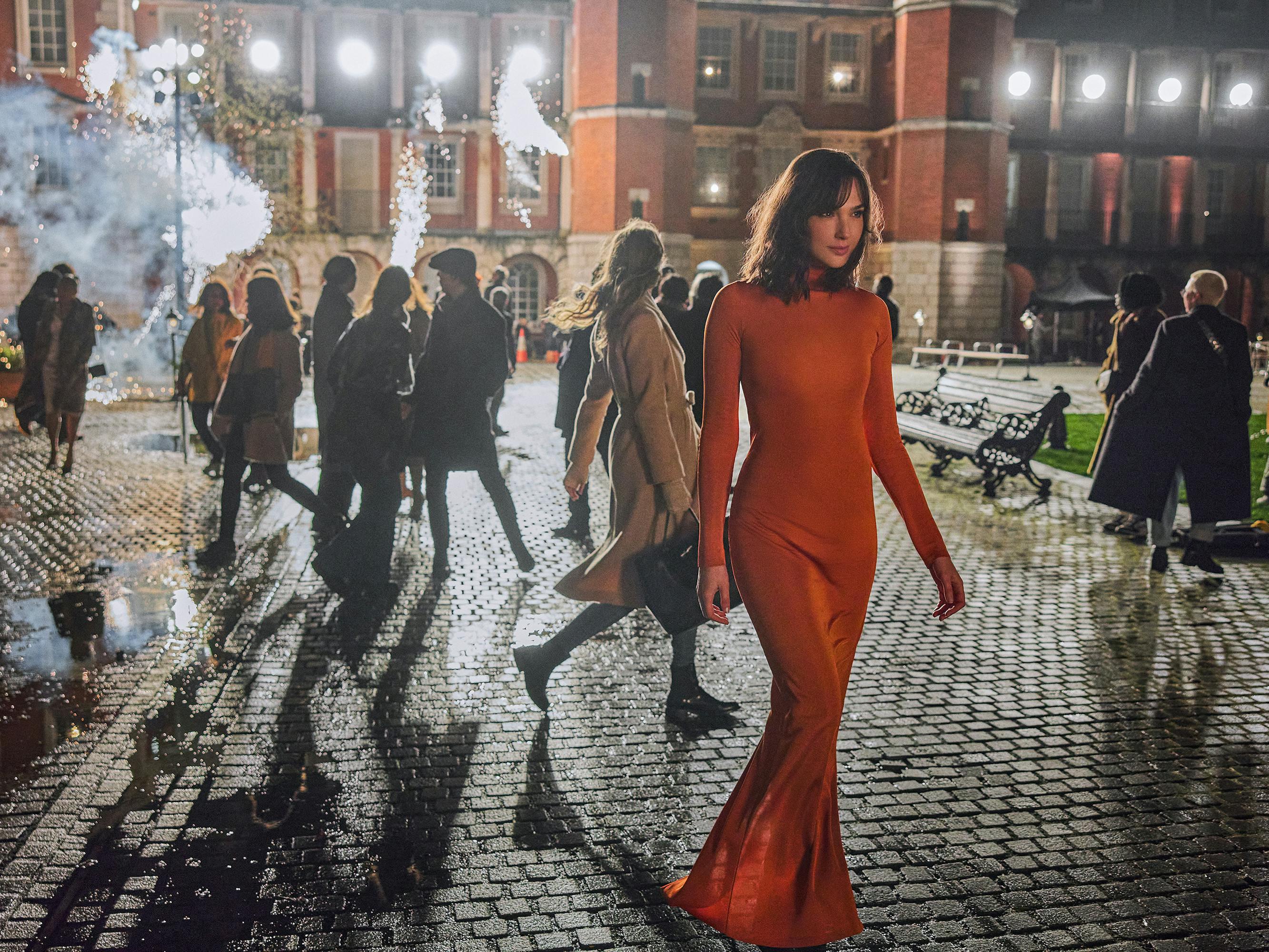 Gal Gadot wears an orange long-sleeved dress and walks through a dark, cobble-stoned street.