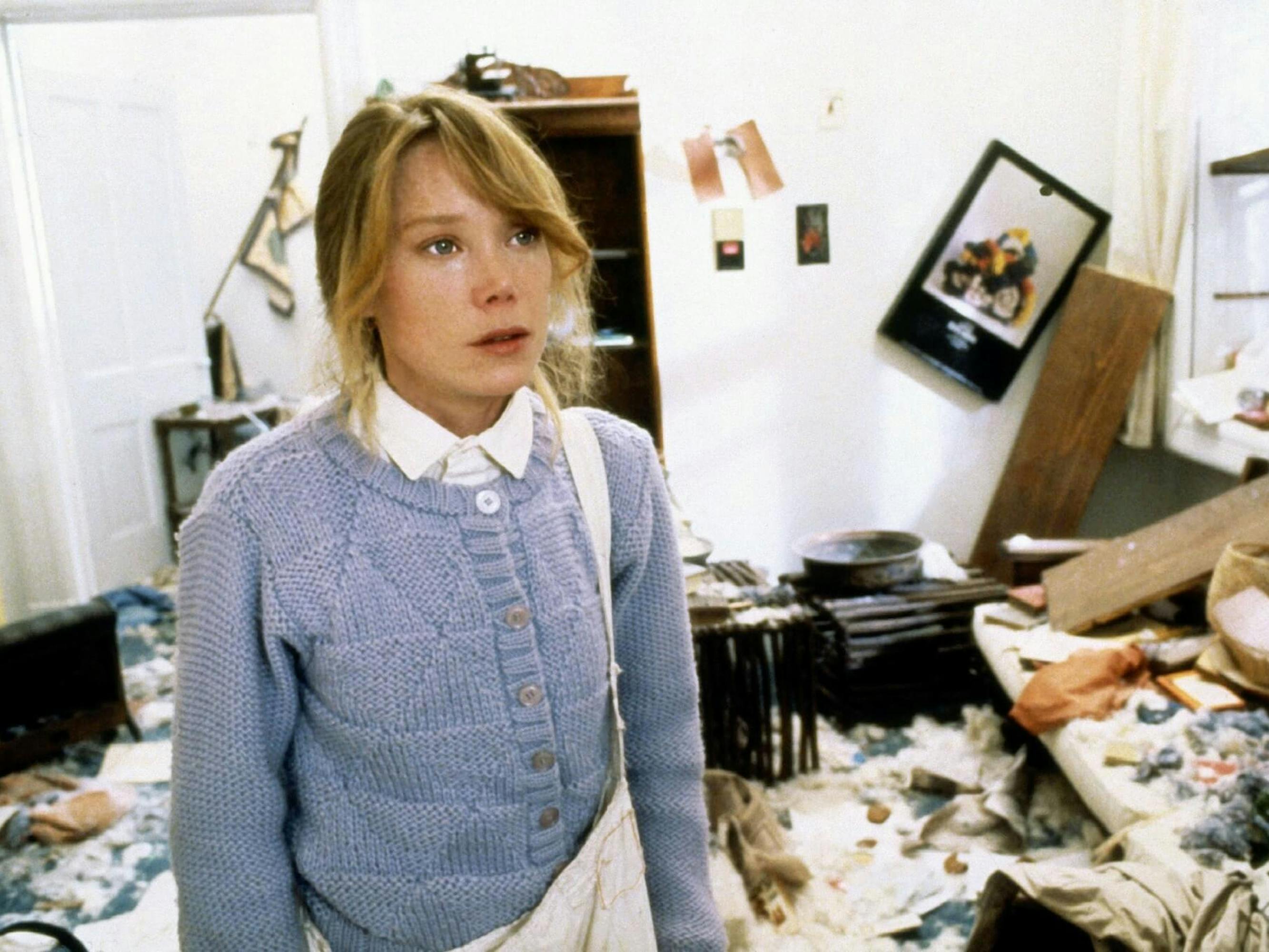 Beth Horman (Sissy Spacek) in Missing tands in a dischevled room with tears in her eyes. She wears a blue cardigan and white collared shirt.
