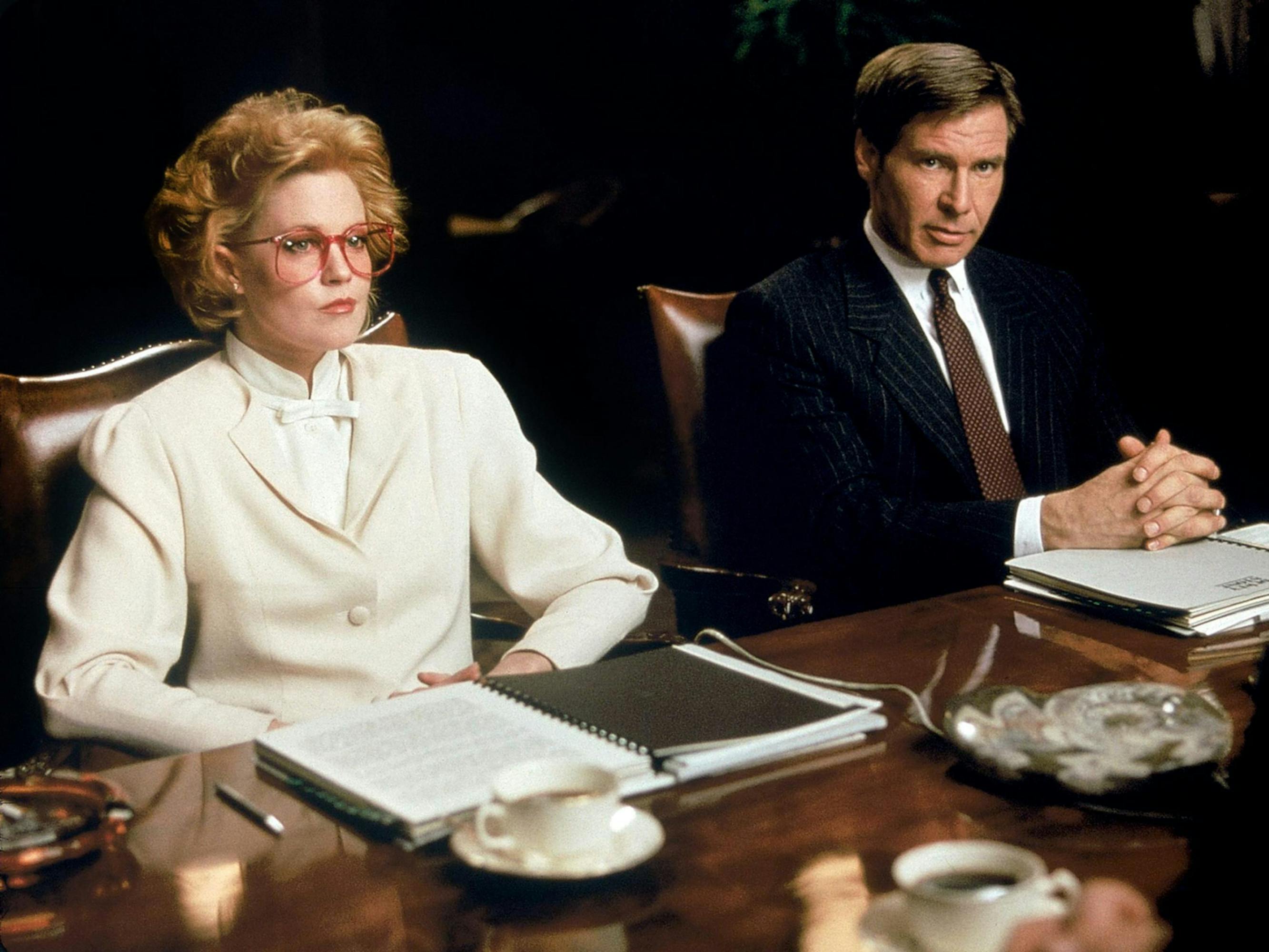 Melanie Griffith and Harrison Ford sit at a table with paperwork sitting in front of them. Griffith has on an all-white suit and red-framed glasses. 