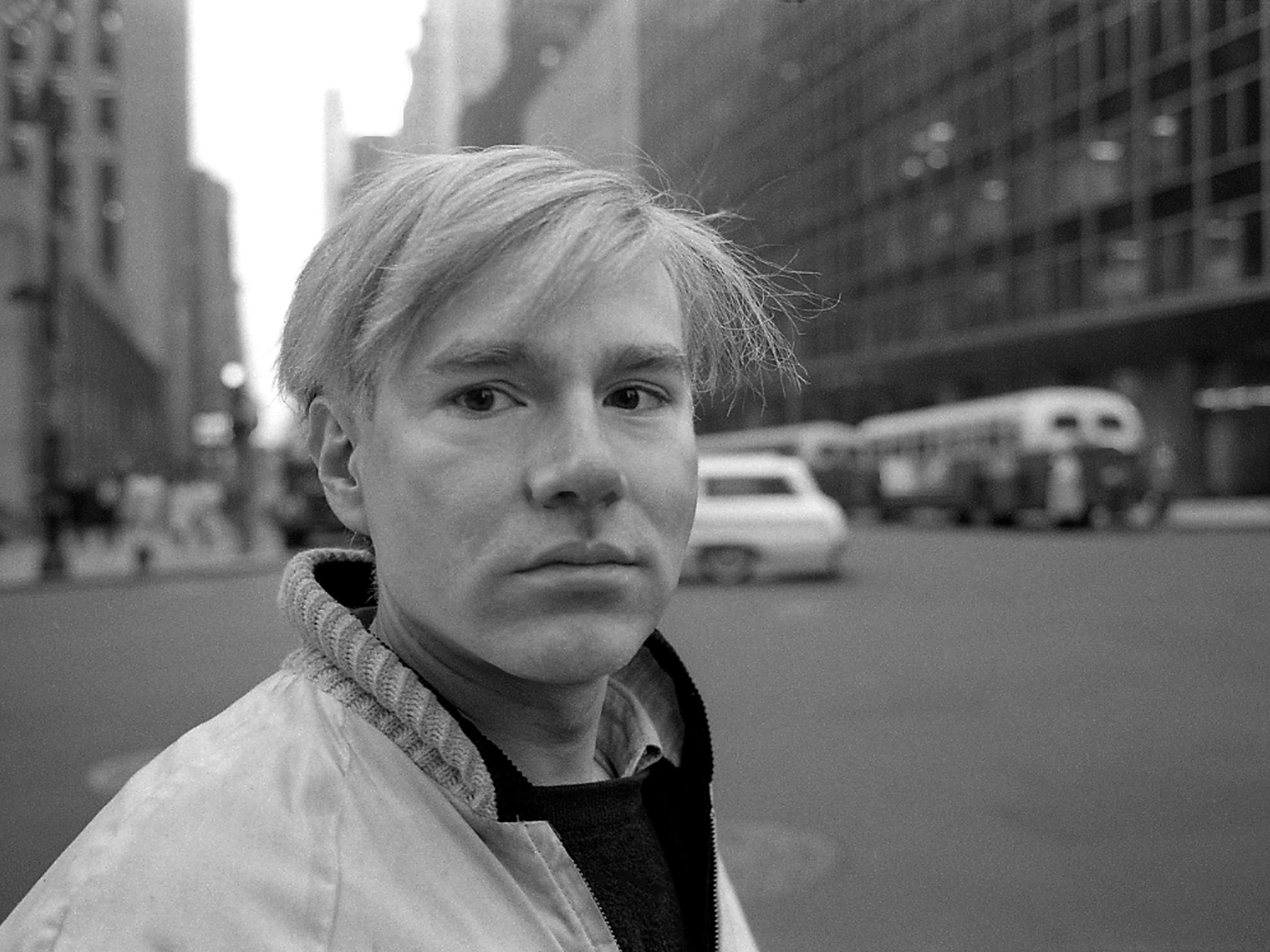Andy Warhol wears a light colored jacket and stands in the middle of the street.