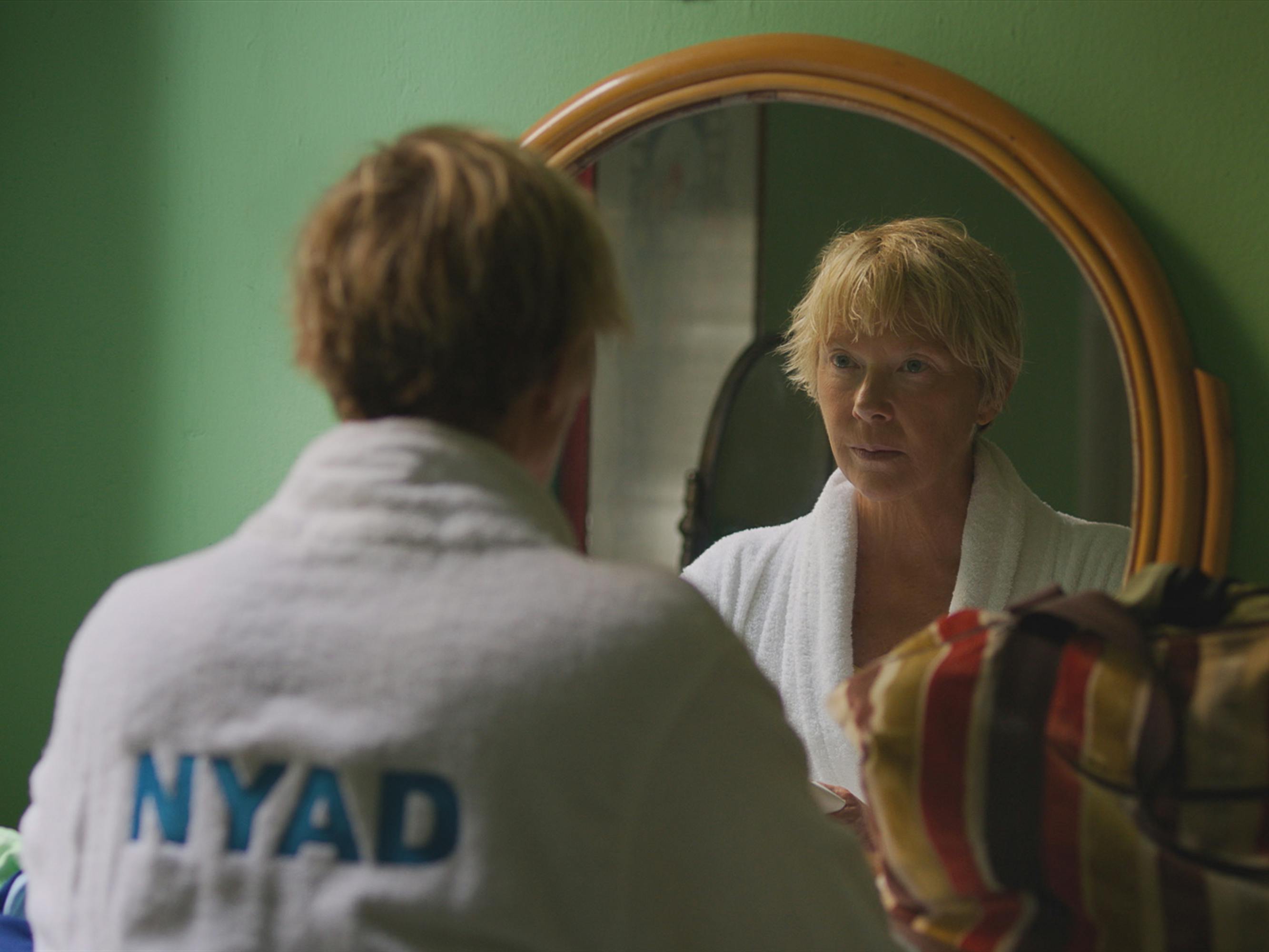 Diana Nyad (Annette Bening) wears a white robe with NYAD in blue writing on the back and looks into the mirror.