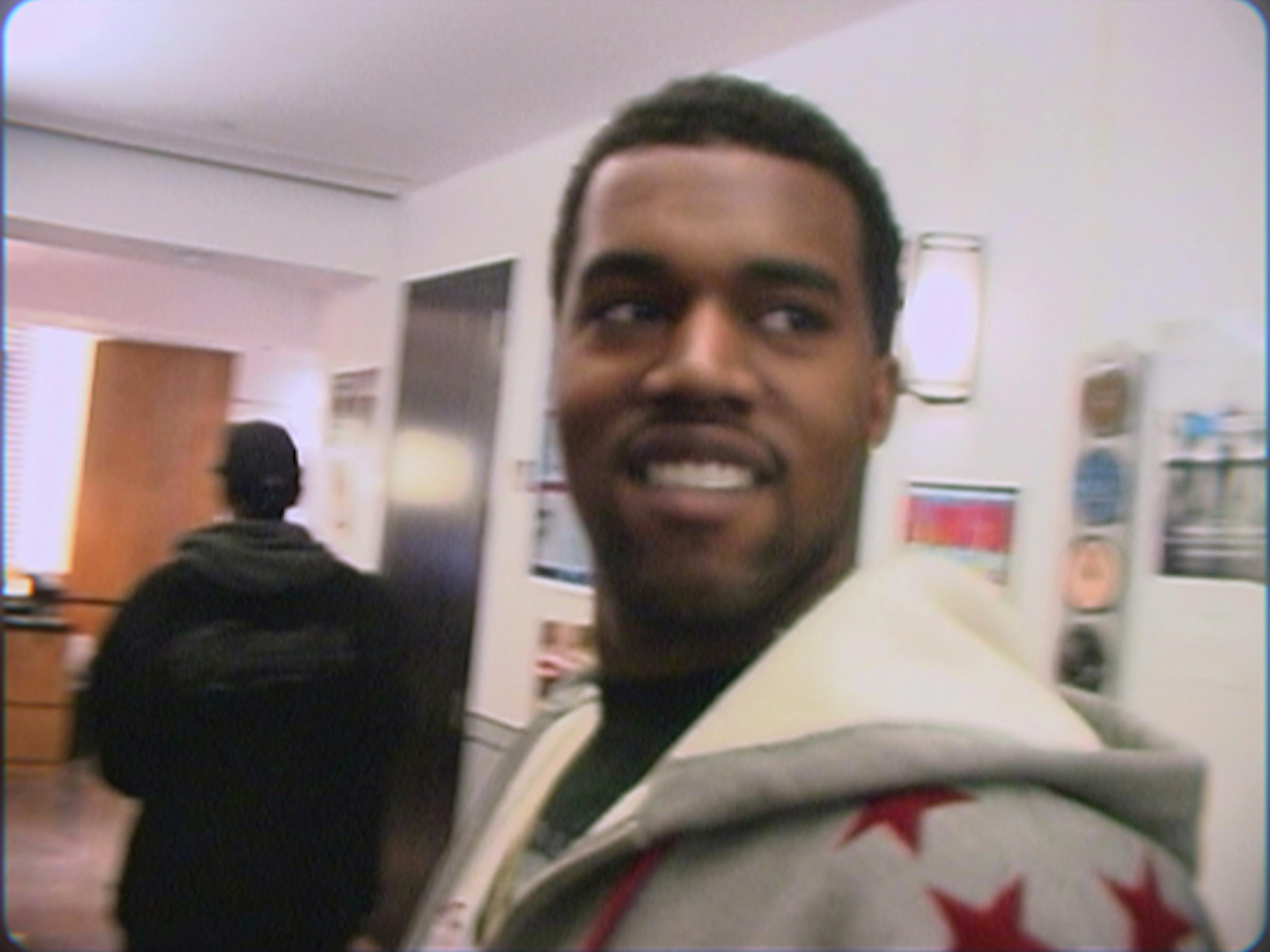Kanye West wears a grey hoodie with red stars and smiles in this close up picture.