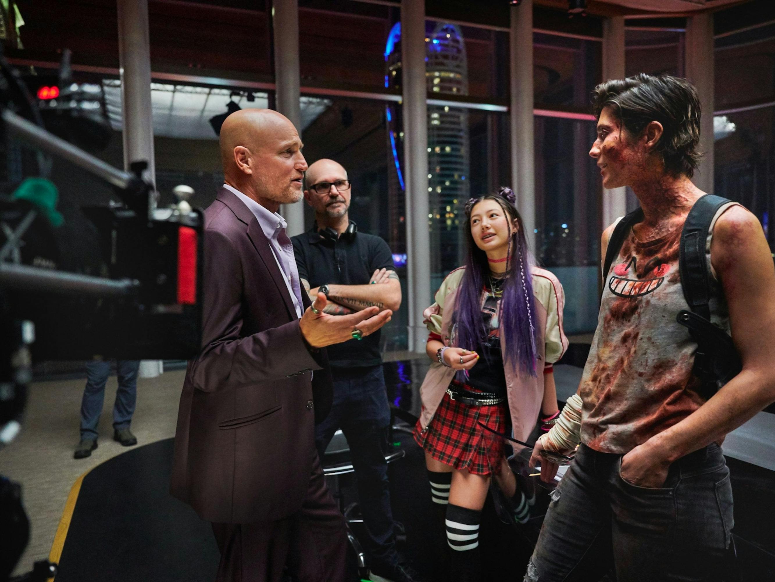 Woody Harrelson, Director Cedric Nicolas-Troyan, Miku Martineau and Mary Elizabeth Winstead stand in a half circle listening to Woody explain something with his hands. The room is walled with floor to ceiling windows, and there is camera gear and camera people mingling about.