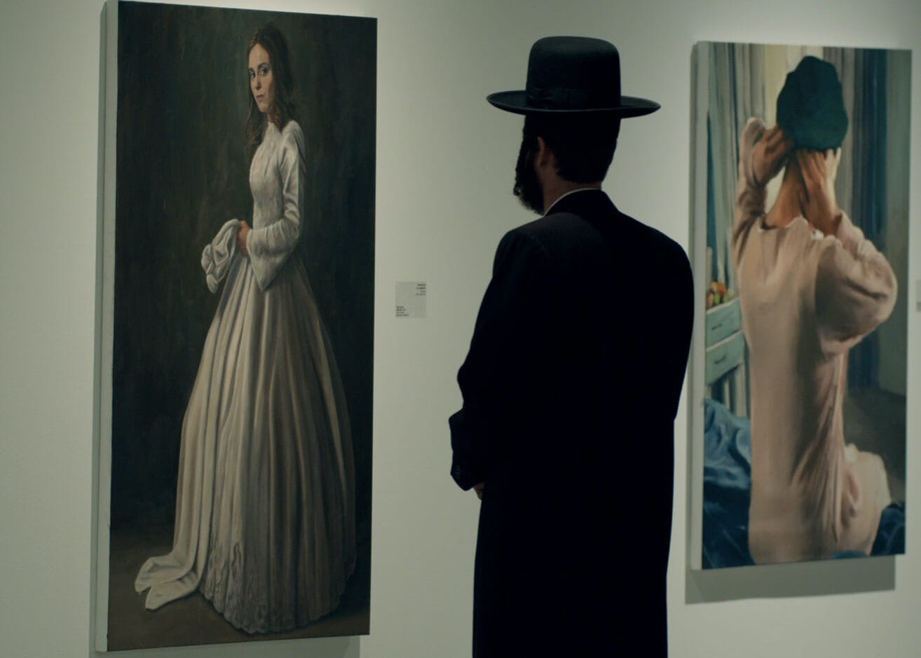 A man stares at two paintings. He wears all black, including a black hat. The painting closer to the man is a woman in a long white dress against a black background. The further painting is a person in white sitting on a blue bed adjusting their hair. There is a turquoise piece of furniture in the background.