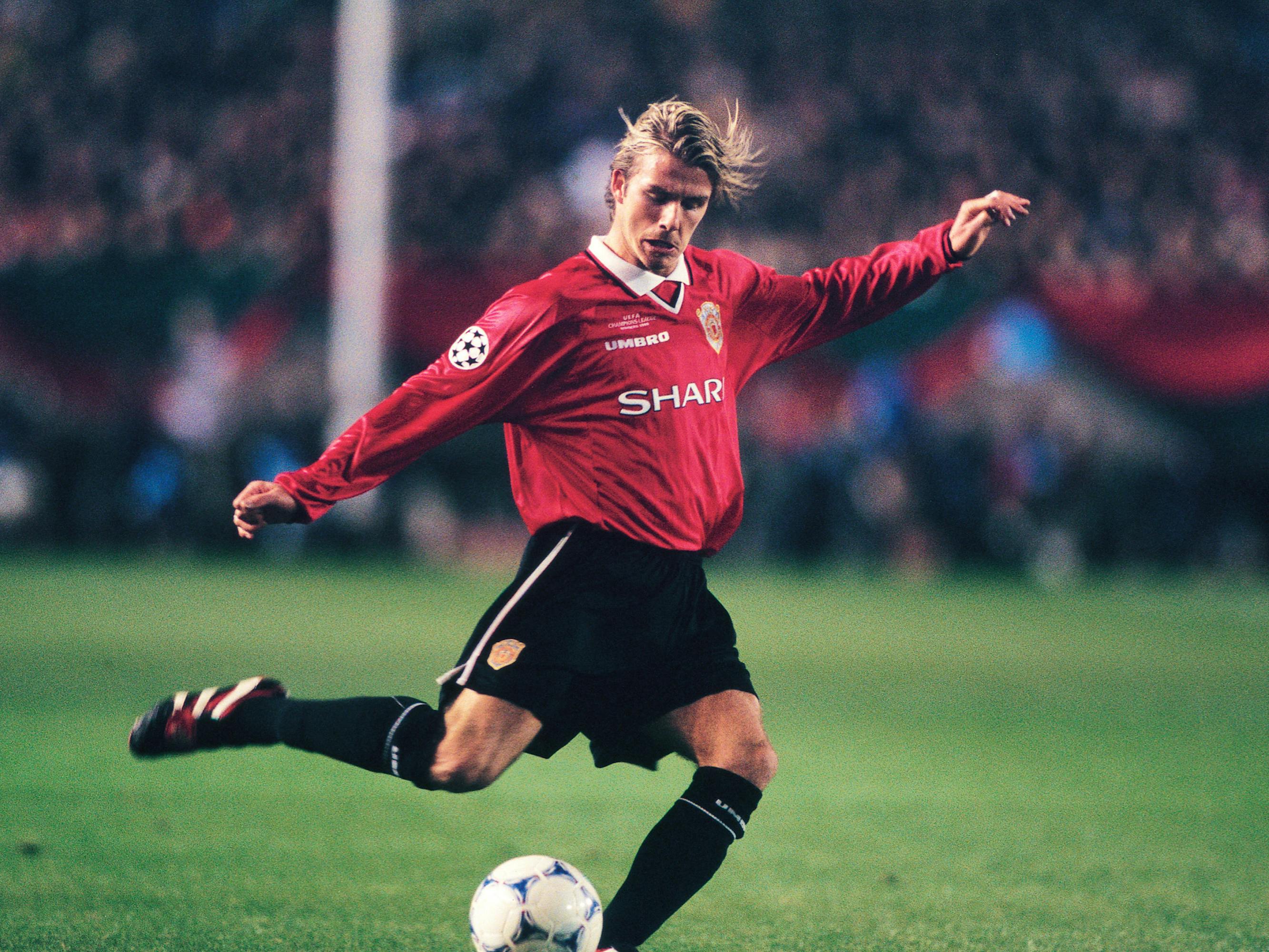 David Beckham wears a red jersey and black shorts and kicks a ball on a green soccer pitch.