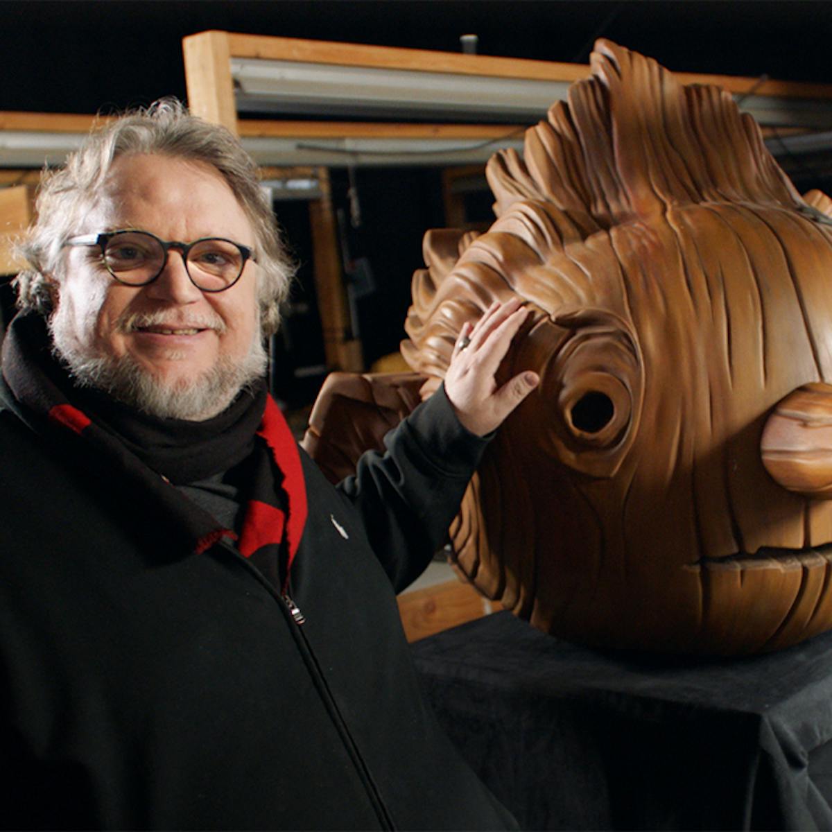 Guillermo del Toro stands next to Pinocchio’s head. Del Toro wears a black jacket and a black-and-red scarf. 
