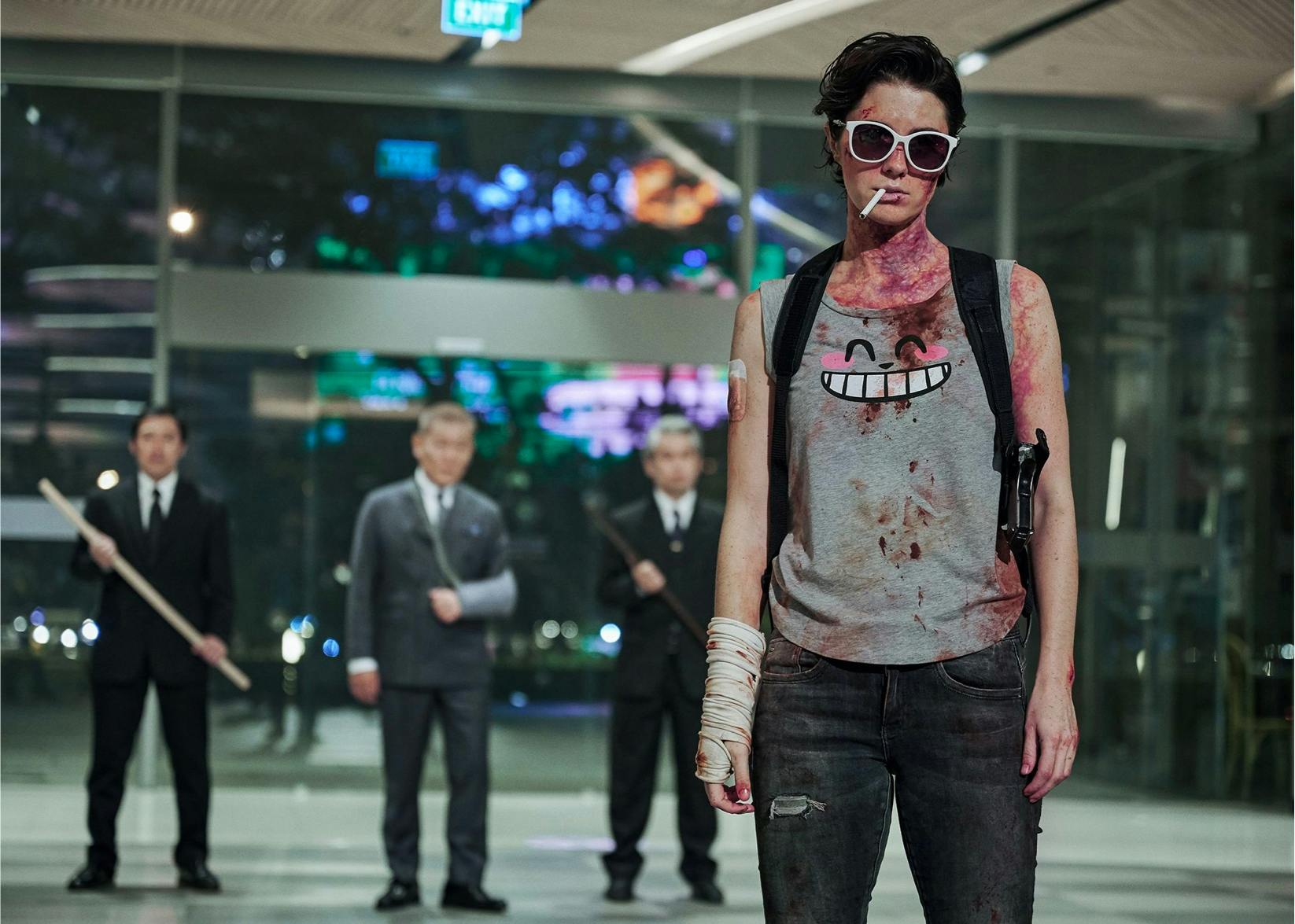 A bruised and bloodied Kate (Mary Elizabeth Winstead) stands in a building lobby with three men standing at a distance behind her.  She wears white-rimmed sunglasses, a grey vest with a smiling face on it, jeans, a shouldered gun holster, and a bandage on her right wrist.  A cigarette hangs nonchalantly from the corner of her mouth. One of the three men has his left arm in a sling, and the men on either side of him are armed with long batons.