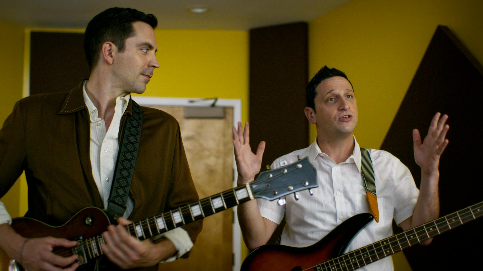  Lead singer (Rhys Coiro) and bass player (Tim Robinson) sing in a yellow walled room.