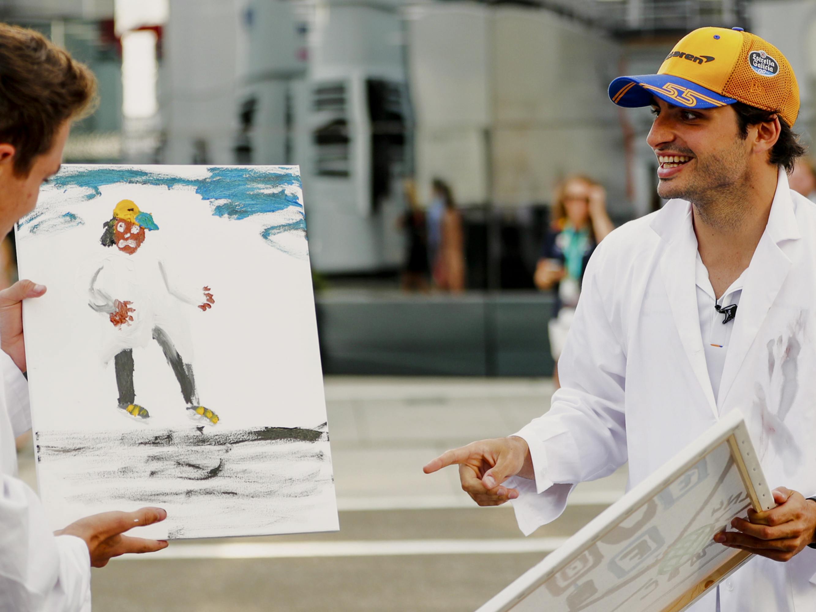 Lando Norris stands on the left holding a painting he has made of friend and teammate Carlos Sainz Jr. Sainz Jr. stands on the right, pointing to the painting and smiling large. 